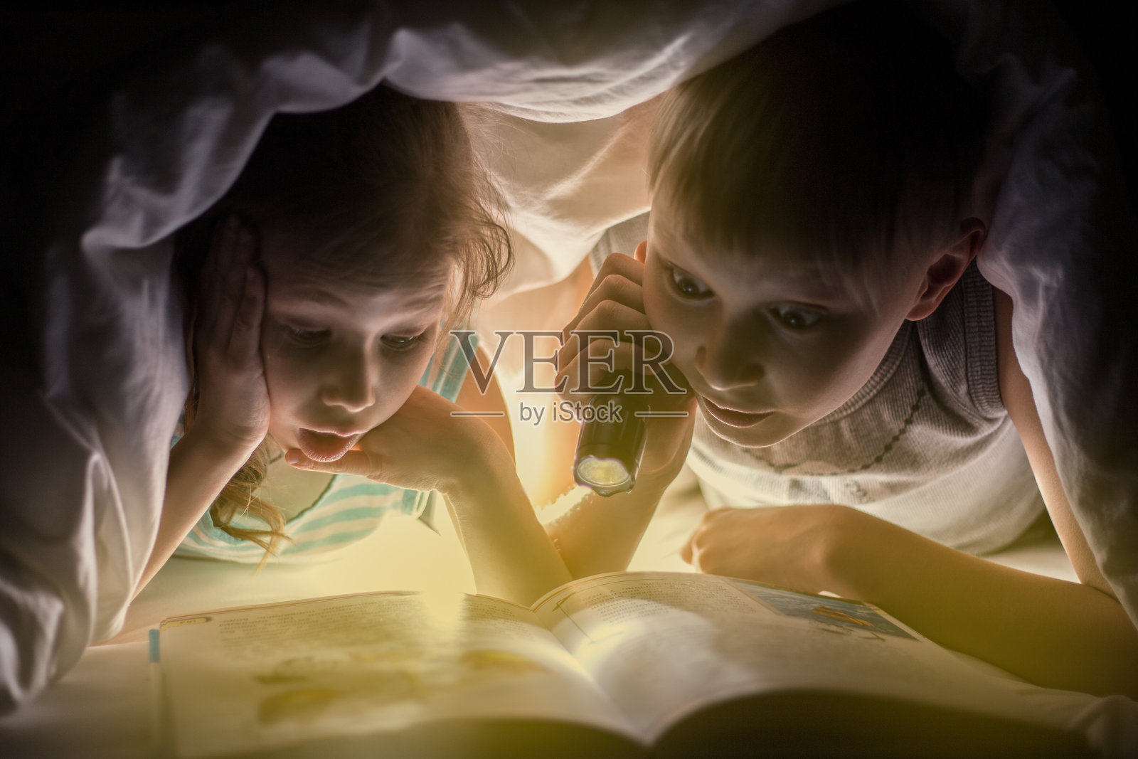孩子睡觉。妹妹和弟弟正在用手电筒盖着毯子看书。漂亮的年轻男孩和可爱的女孩有乐趣在儿童房。照片摄影图片