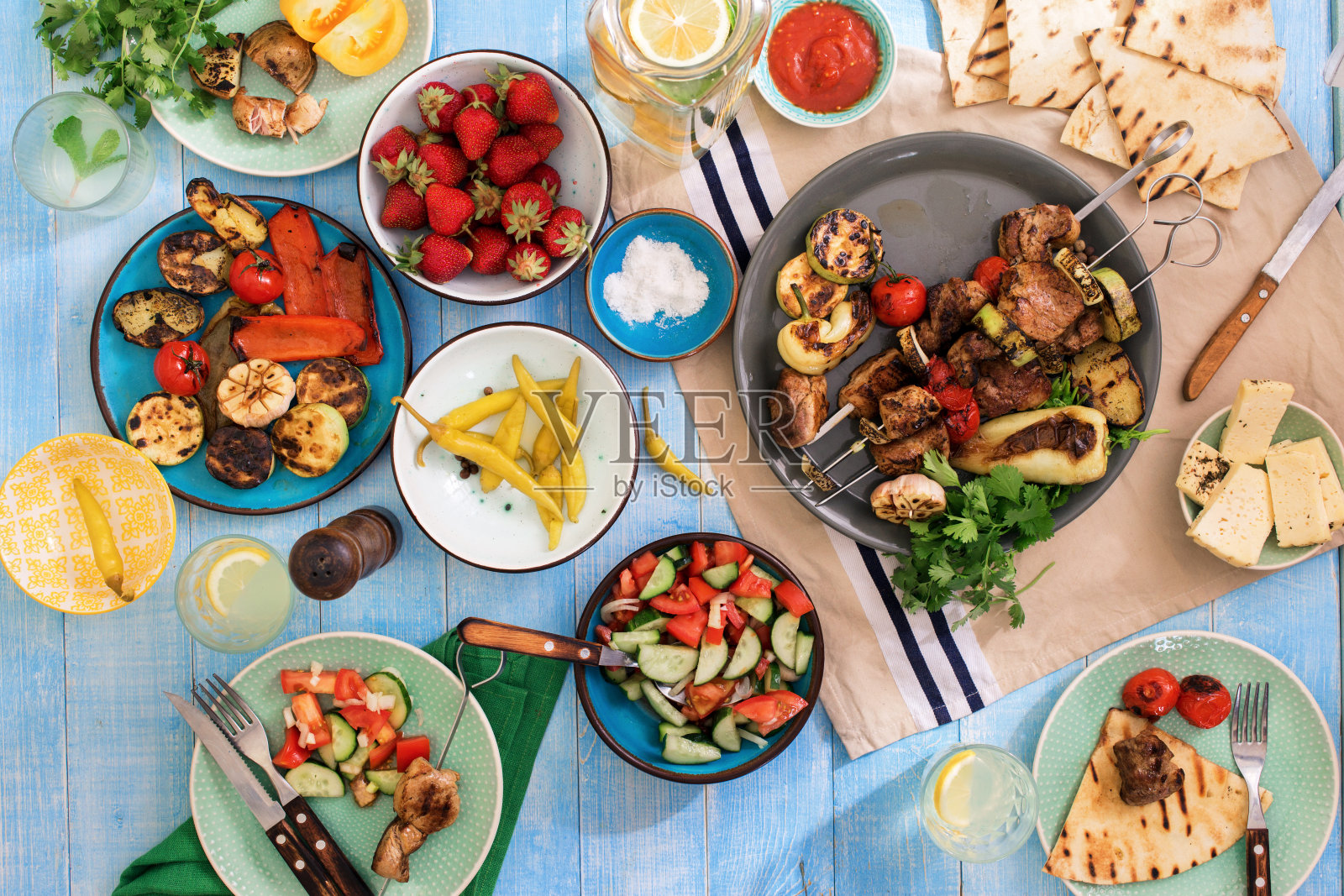 蓝色的木桌上放着烤肉串、各种烤蔬菜、沙拉、柠檬水、草莓和零食照片摄影图片