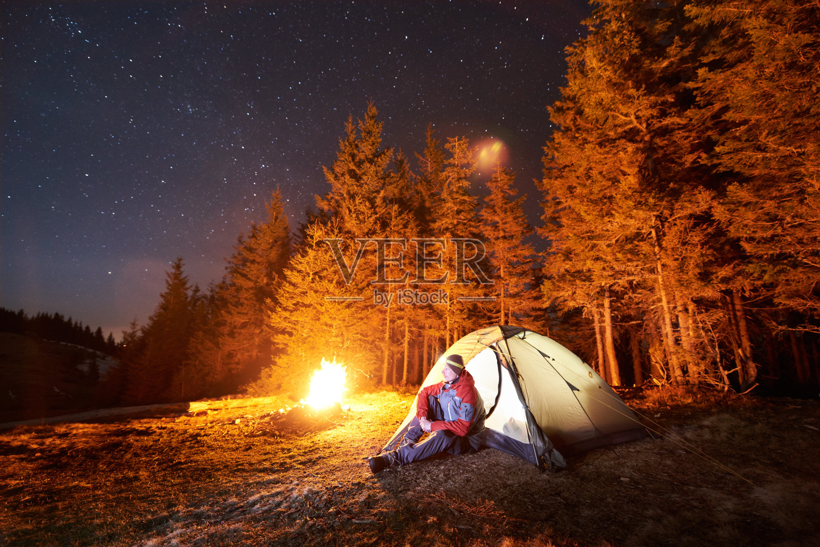 男游客晚上在森林附近的营地休息。一个人坐在篝火和帐篷附近，在美丽的夜空下满是星星，欣赏夜景照片摄影图片