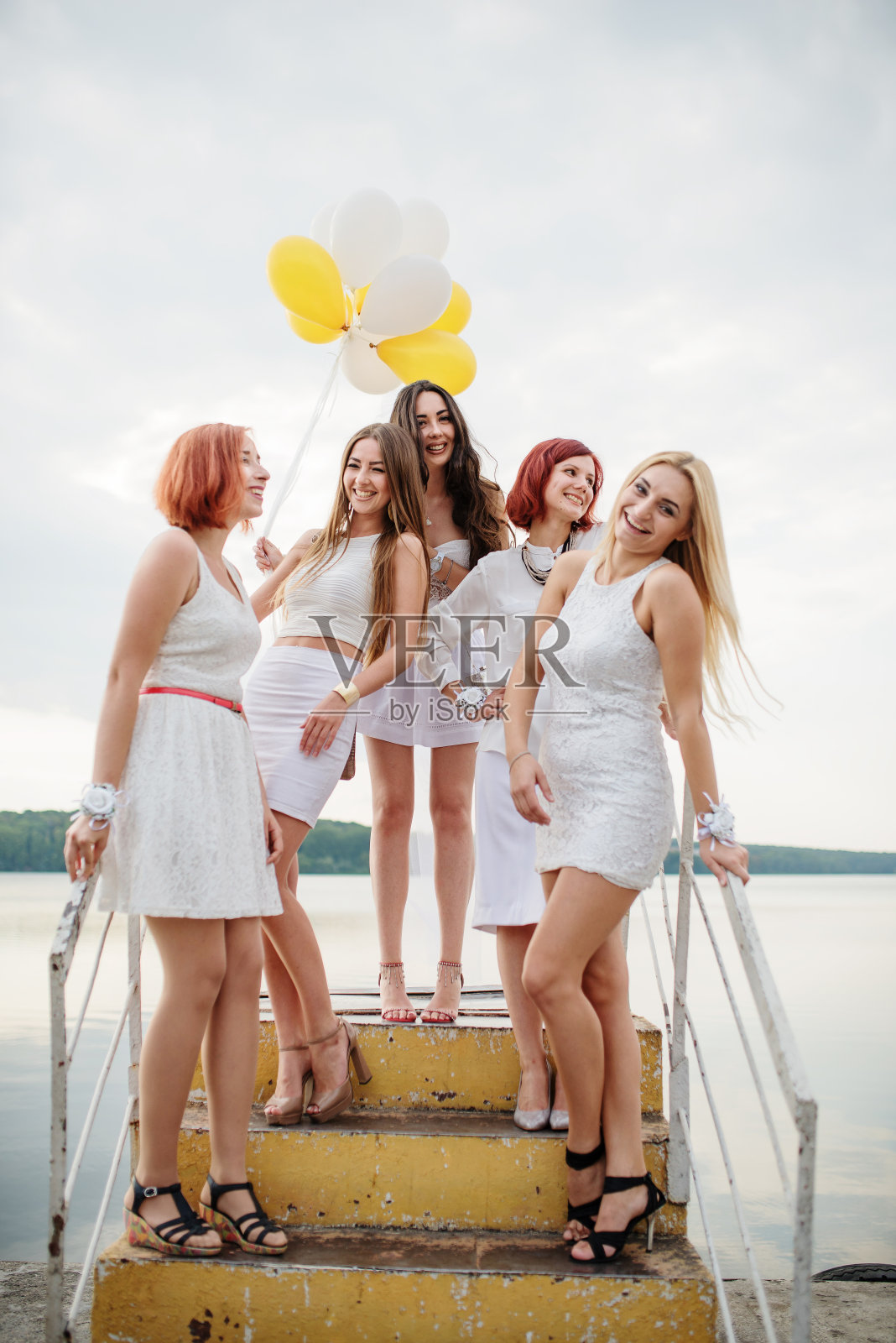 五个手里拿着气球的女孩穿着白色的衣服，在湖边的码头上参加女性聚会。照片摄影图片