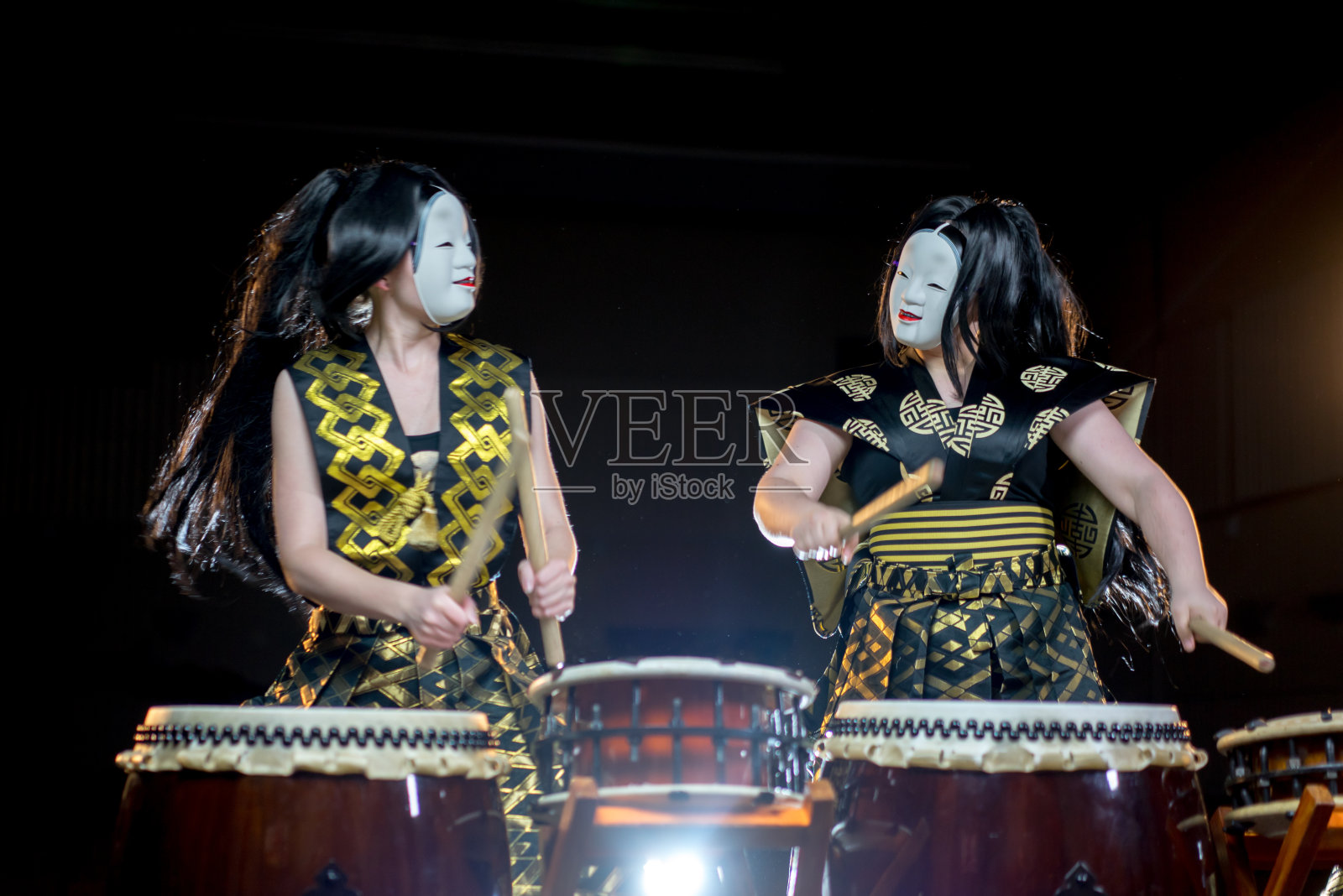 两个鼓手女孩在白色的恶魔面具和鼓槌，工作室拍摄在一个黑暗的背景。照片摄影图片