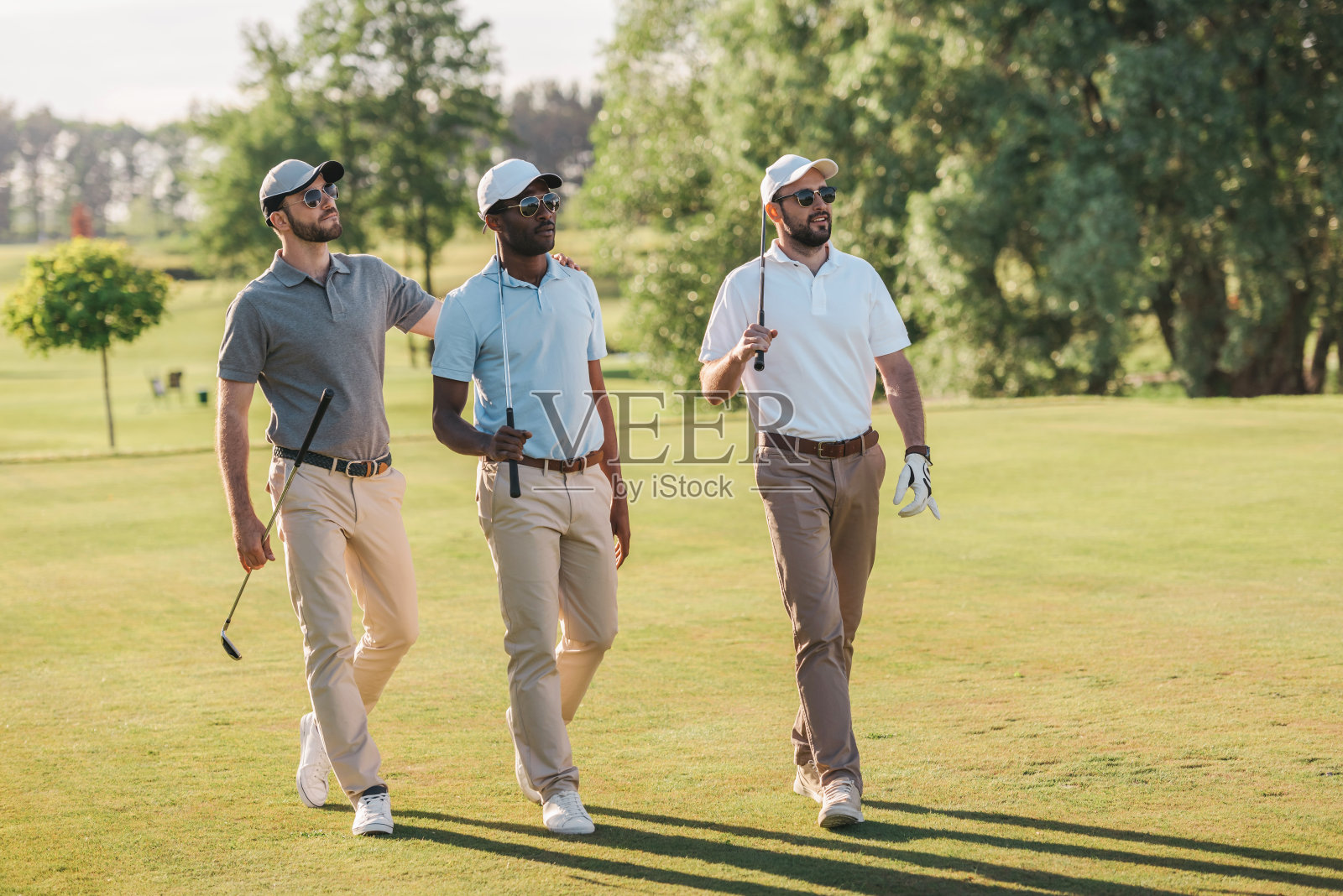 戴着帽子和太阳镜的自信男人拿着高尔夫球杆在草坪上走着照片摄影图片