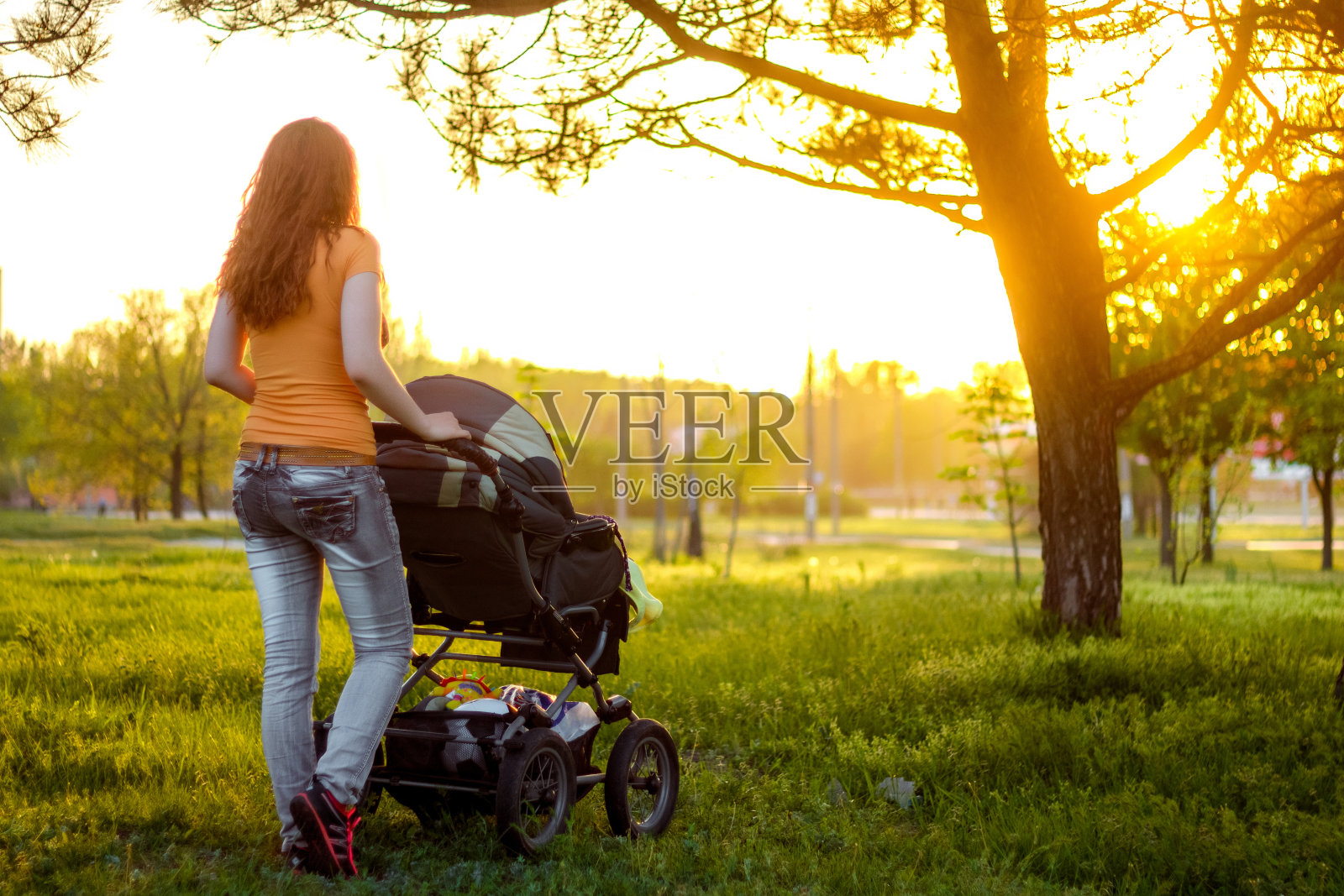 年轻的妈妈推着婴儿车在公园里散步，从后面看，傍晚的阳光普照照片摄影图片