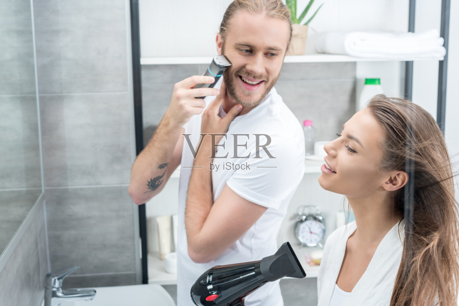 英俊的年轻人用电动剃须刀刮胡子，微笑的女人用吹风机吹头发照片摄影图片