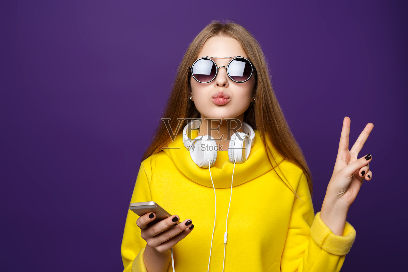 描绘年轻的少女戴着耳机和电话，穿着黄色毛衣，孤立在紫色的背景上。照片摄影图片