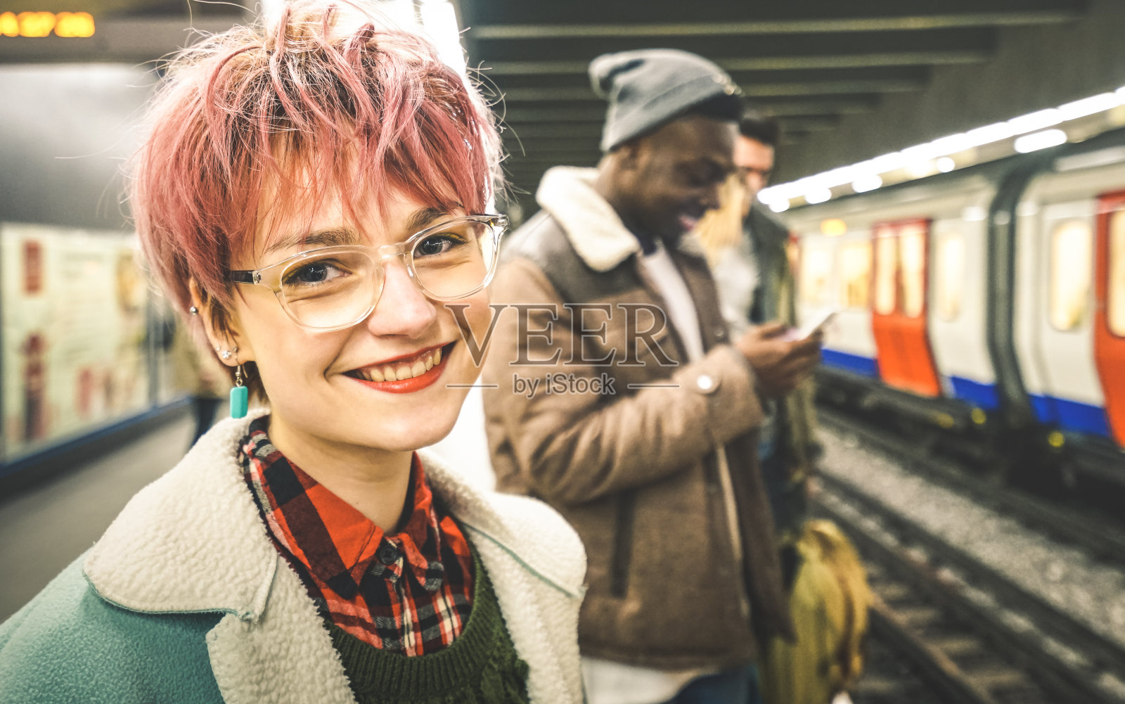 粉红色头发的年轻女子和一群多种族的潮人朋友在地铁车站等火车-城市友谊的概念与年轻人一起站在城市地下区域照片摄影图片