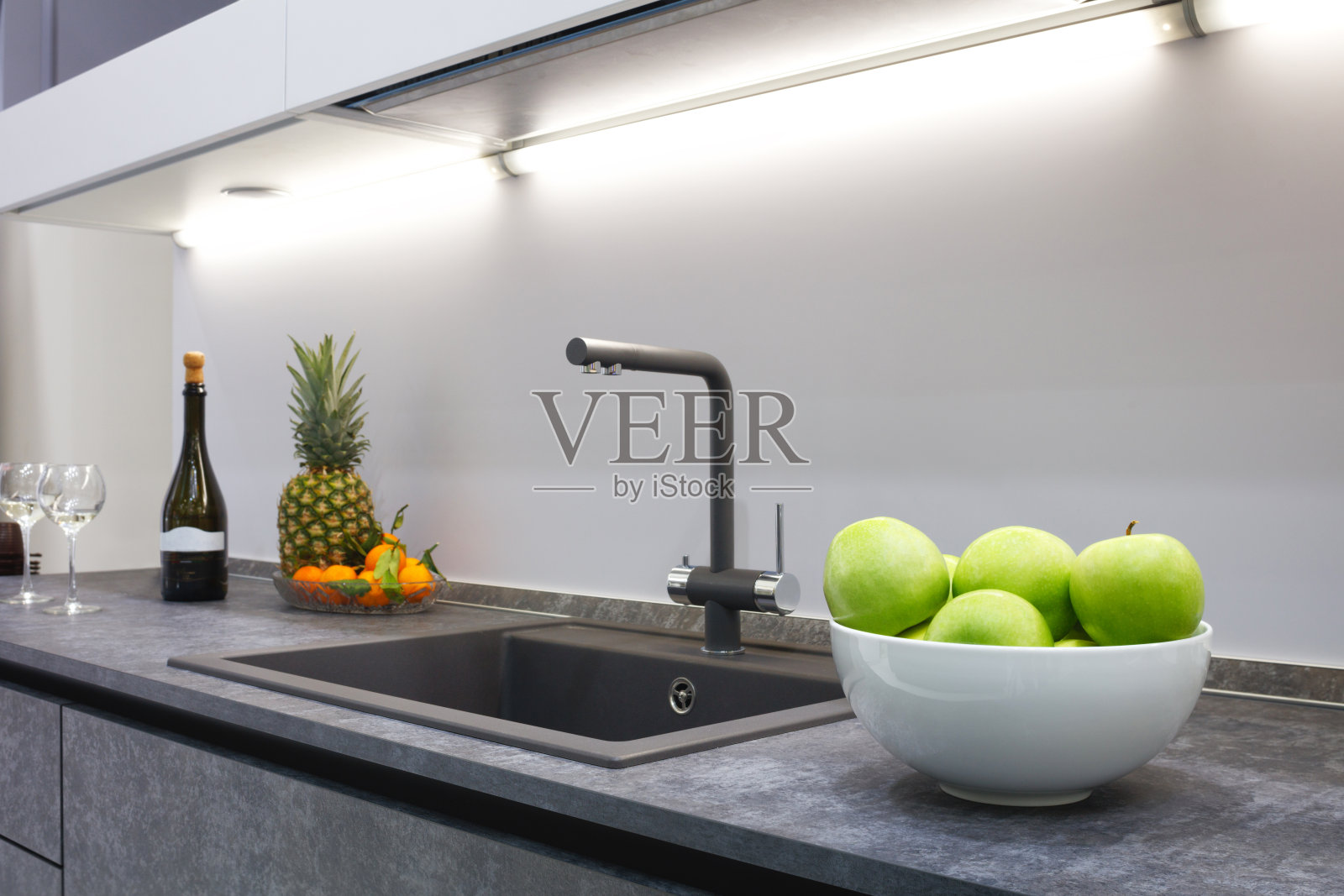 这间现代厨房的内部装饰着一个灰色的石材台面，里面有一个豪华的洗脸盆和搅拌机、菠萝和橘子、一瓶红酒和两个玻璃杯。照片摄影图片