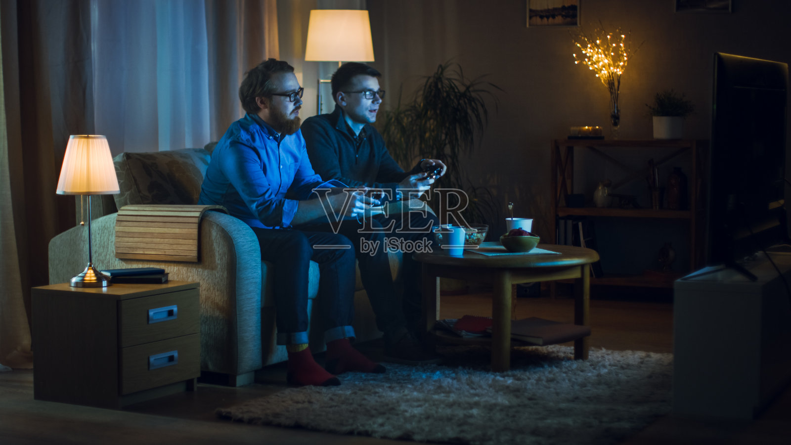 晚上两个朋友坐在客厅的沙发上玩电子竞技游戏。他们互相推搡，很友好。照片摄影图片