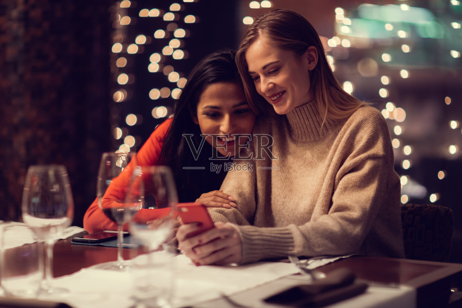 两个年轻的女人在一家高级餐厅里走过菜单，一边喝着红酒，一边有说有笑，也许还在闲聊，一边一直看自己的手机照片摄影图片