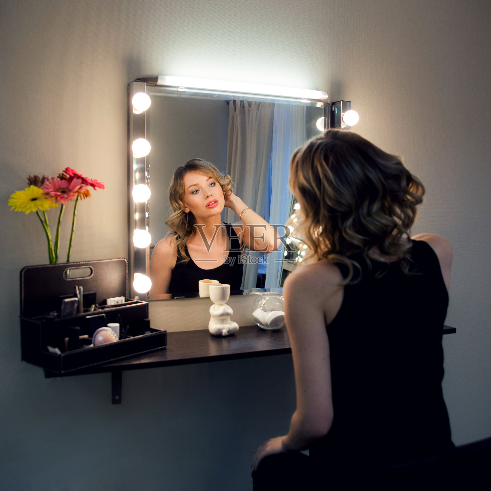 漂亮的金发女郎在美容室里用灯照着镜子打扮。一个女人在美容院欣赏自己的倒影。美丽的脸，自然的妆容和奢华的卷发。照片摄影图片