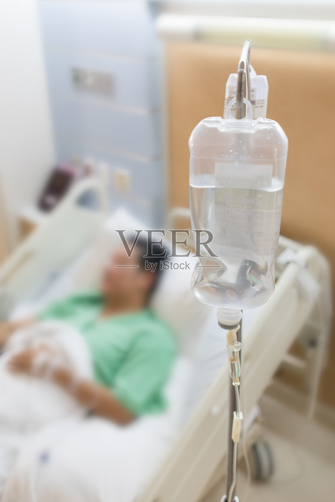 模糊图像的男性病人躺在医院的床上，以盐溶液袋为前景。照片摄影图片