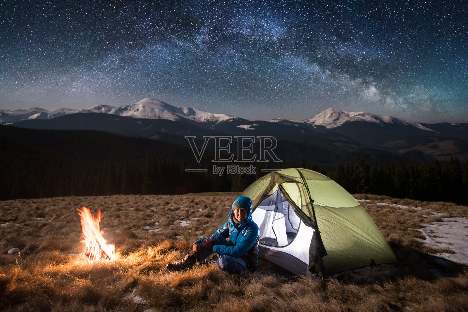 女游客晚上在她的营地。快乐的女人看着镜头，坐在篝火和绿色的帐篷下，在美丽的星空下满是星星和银河。背景是白雪皑皑的群山照片摄影图片