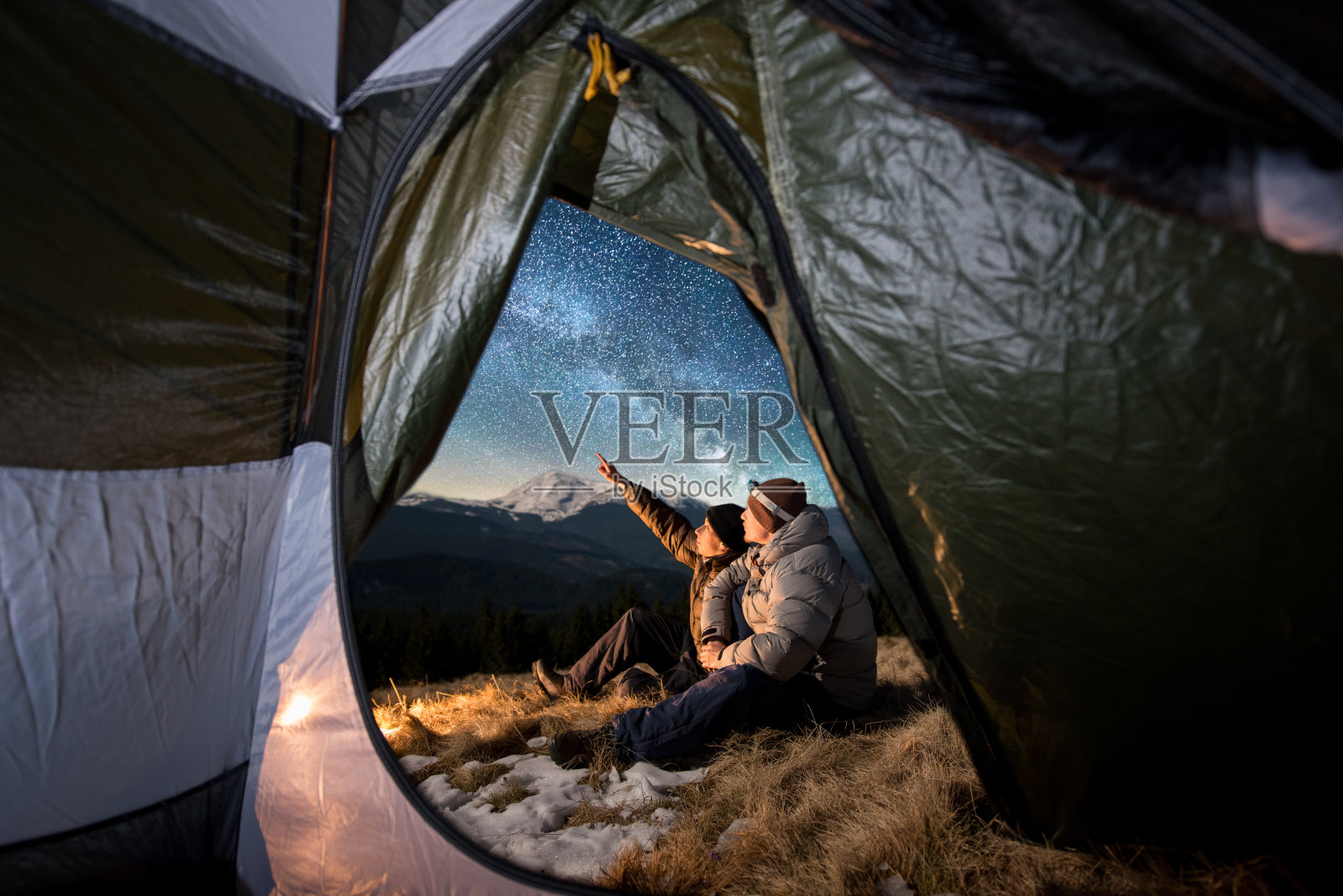从帐篷里面望去，两位游客正在山里的宿营中休息。男人们坐在篝火旁。一个人指着满是星星和银河的美丽夜空照片摄影图片