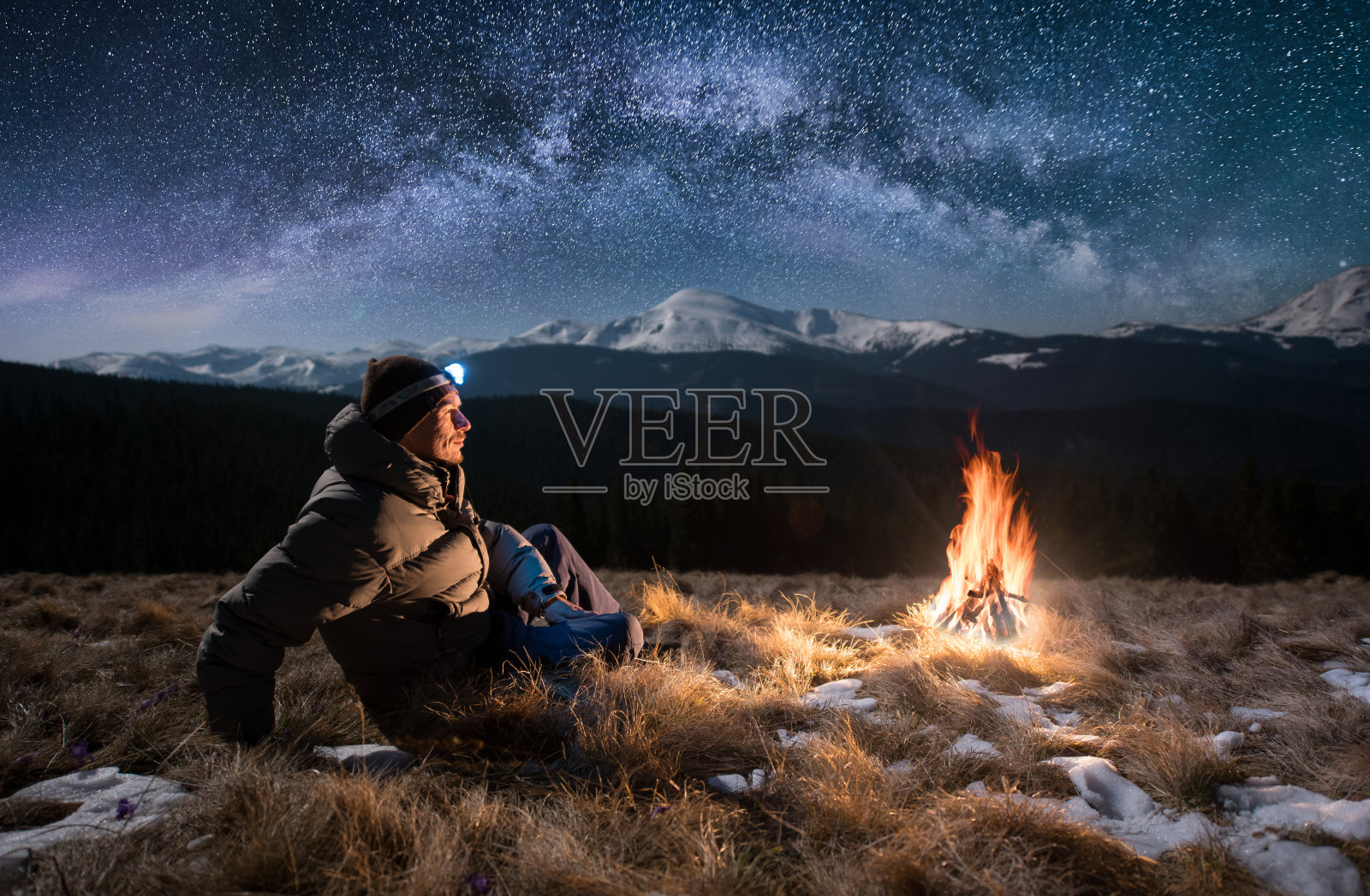 男游客晚上在山上休息。一个人拿着头灯坐在篝火旁，在满是星星和银河的美丽夜空下，欣赏着夜景照片摄影图片