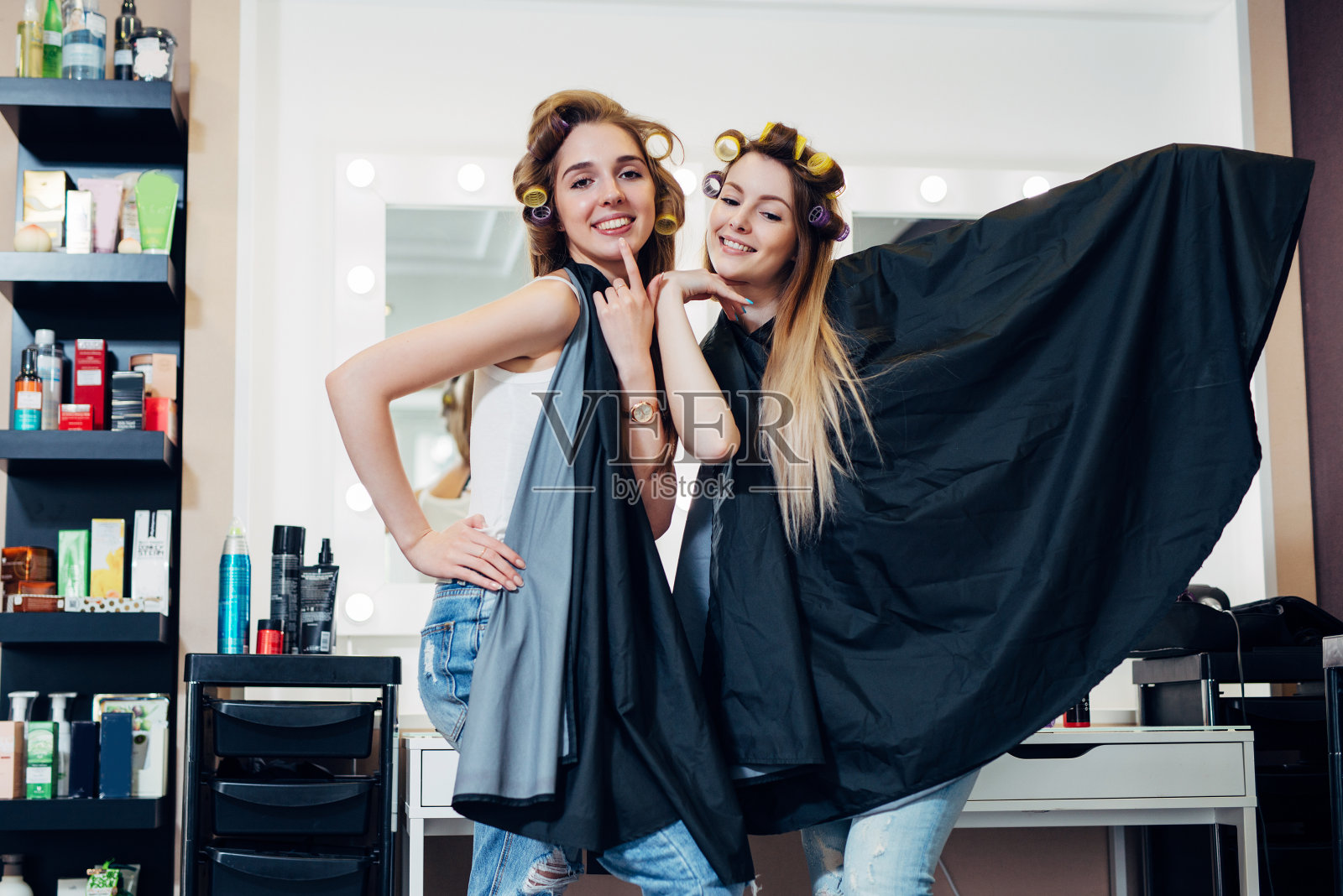 两个漂亮的金发女郎的肖像，戴着卷发器和披肩站在可笑的傻姿势看着相机在美发美容工作室照片摄影图片