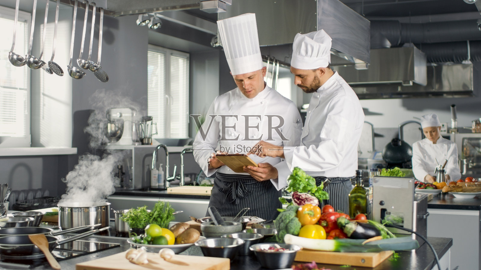两位名厨在平板电脑上讨论他们的视频博客。他们在一个大餐厅不锈钢专业厨房工作。照片摄影图片