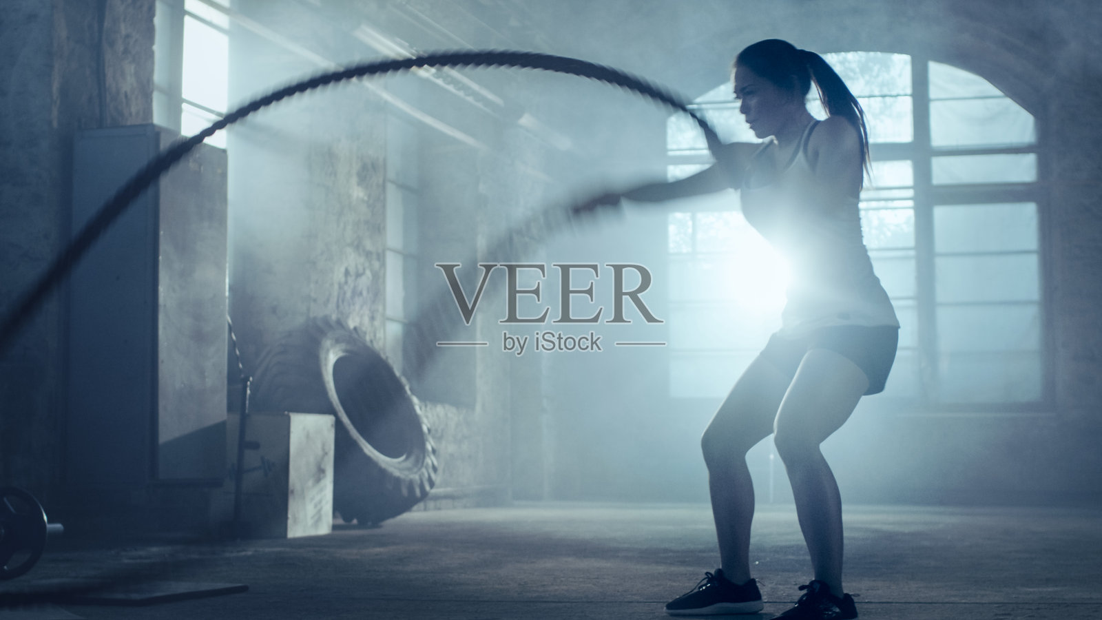 强壮的女运动员练习与战斗绳索作为她的交叉健身健身房锻炼的一部分。她满身的汗水和训练发生在一个废弃的工厂改造成健身房。照片摄影图片