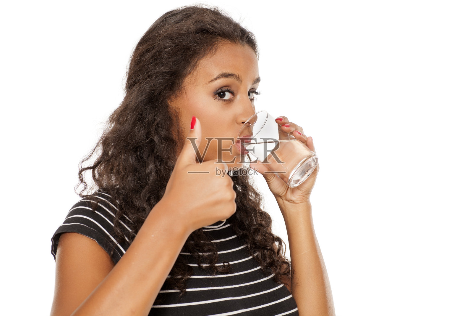 年轻美丽的非洲女孩喝着玻璃杯里的水，在白色的背景上竖起大拇指照片摄影图片