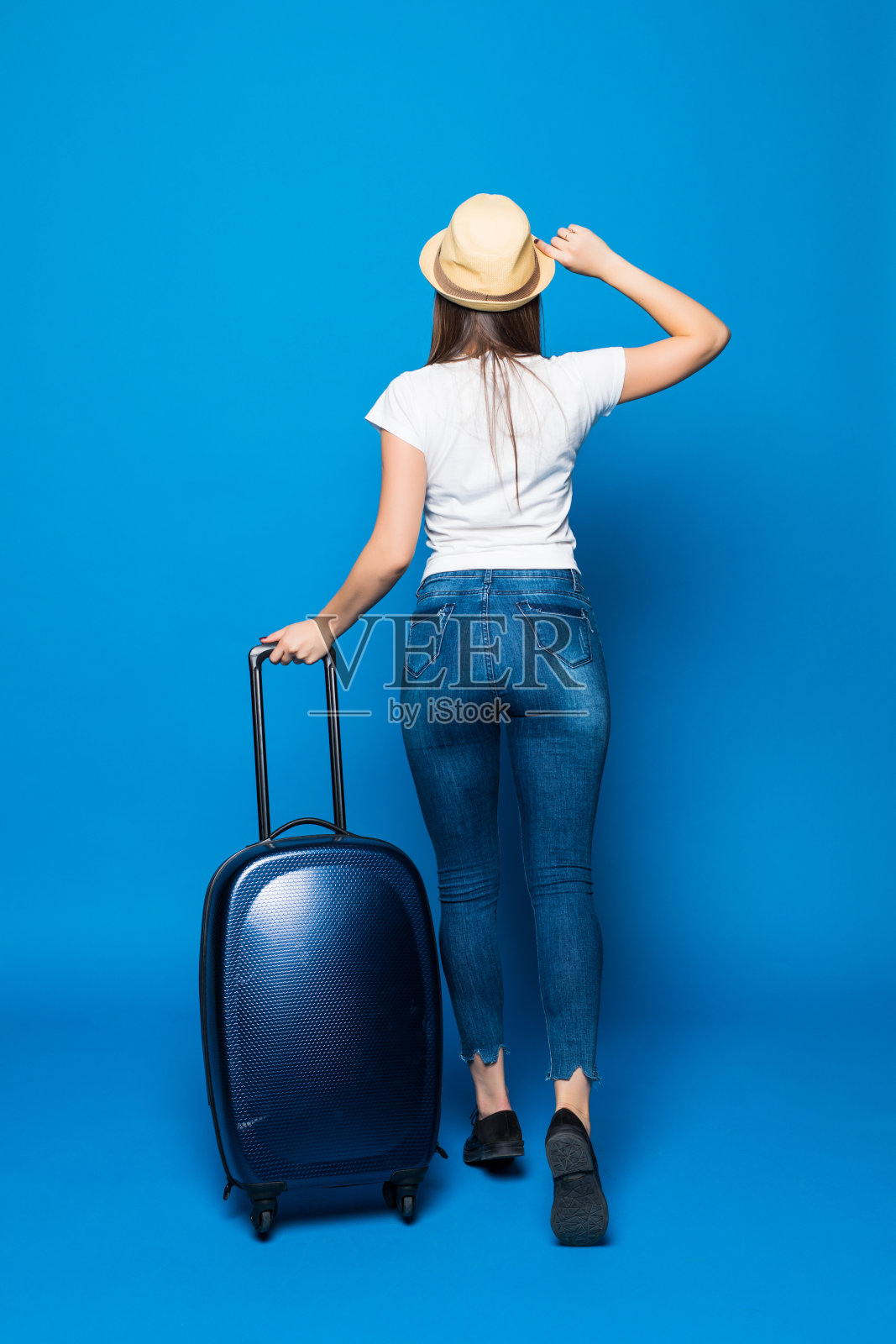 后视图行走的女人与手提箱。美丽的女孩在动。带行李的旅行者的背面视图在蓝色背景上孤立。照片摄影图片