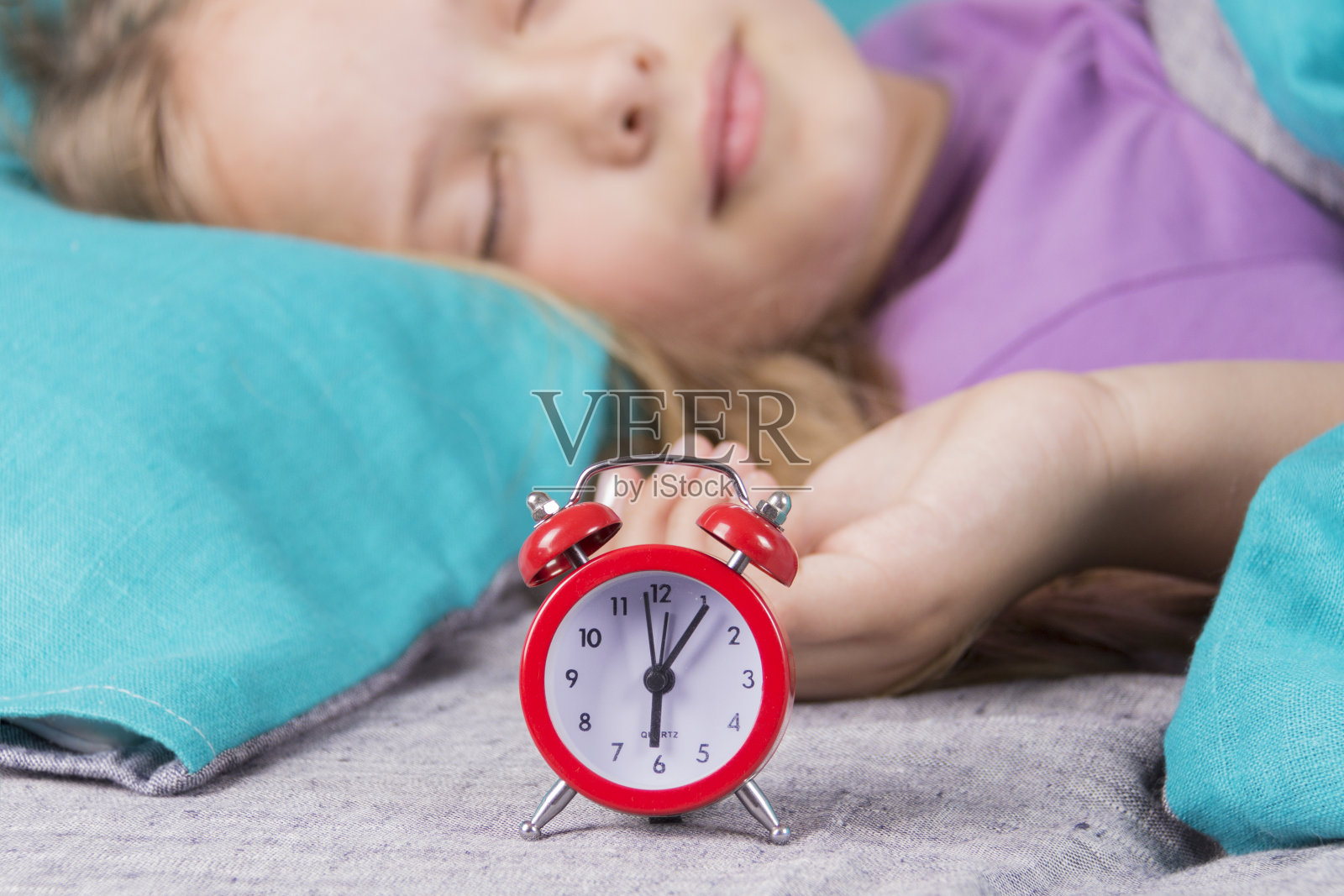 漂亮的小女孩在一个红色闹钟的背景下睡觉，这个闹钟早上会把她吵醒。照片摄影图片