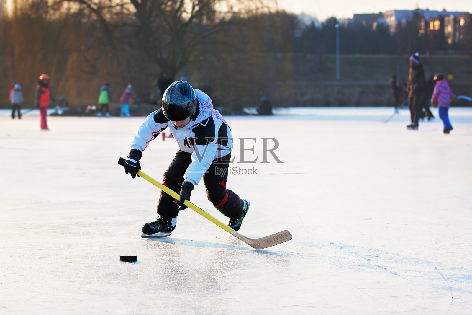 孩子们在公园里冰封的湖面上打曲棍球和滑冰，冬天的日落时分照片摄影图片