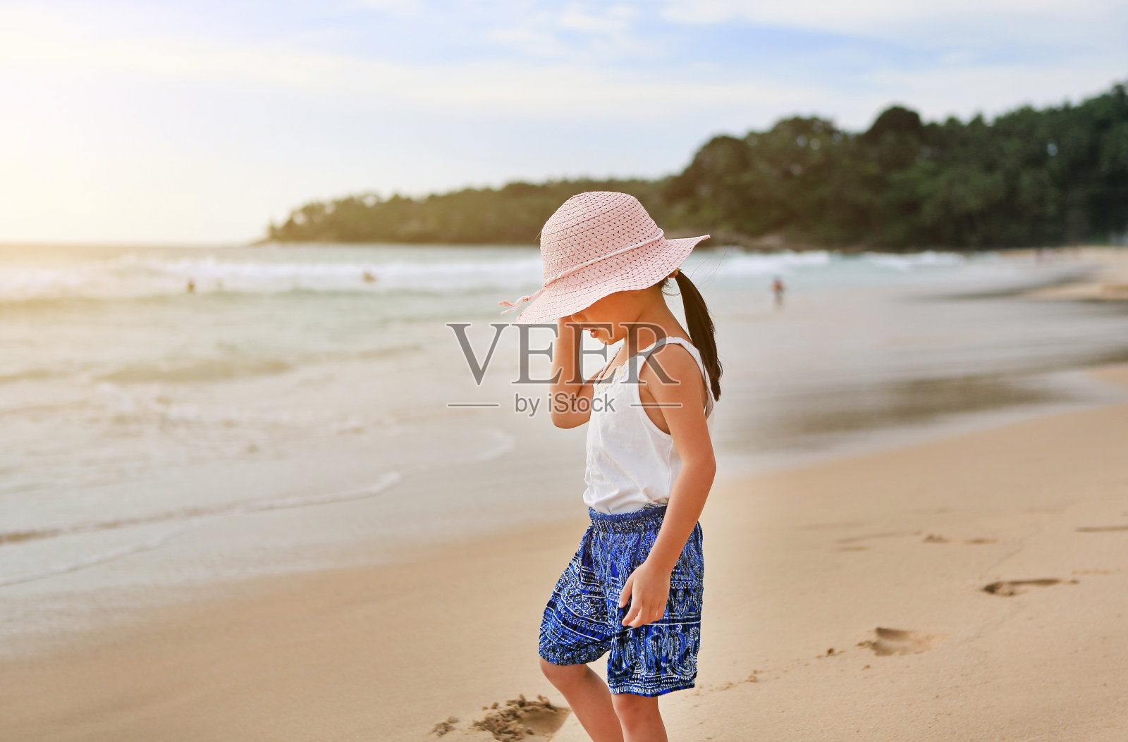 一个亚洲小女孩戴着草帽在海滩上散步。照片摄影图片
