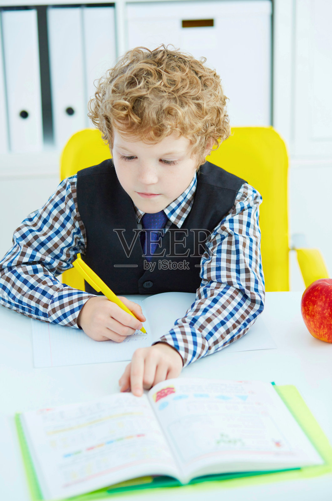 一个白人小男孩坐在桌子旁在笔记本上写字。红苹果挨着桌子。照片摄影图片