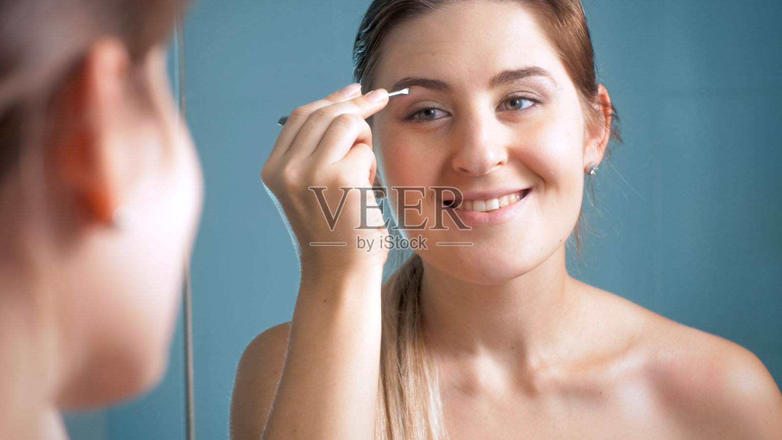 一个快乐微笑的女人在浴室拿着眉钳的肖像照片摄影图片