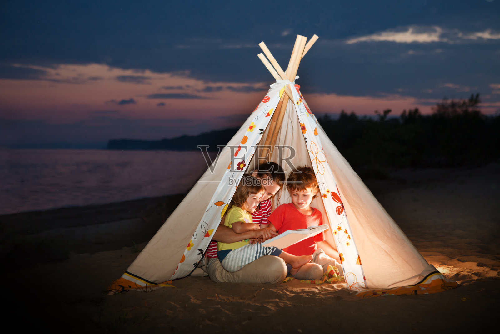 傍晚时分，妈妈和两个孩子在河边的帐篷里用手电筒看书。照片摄影图片