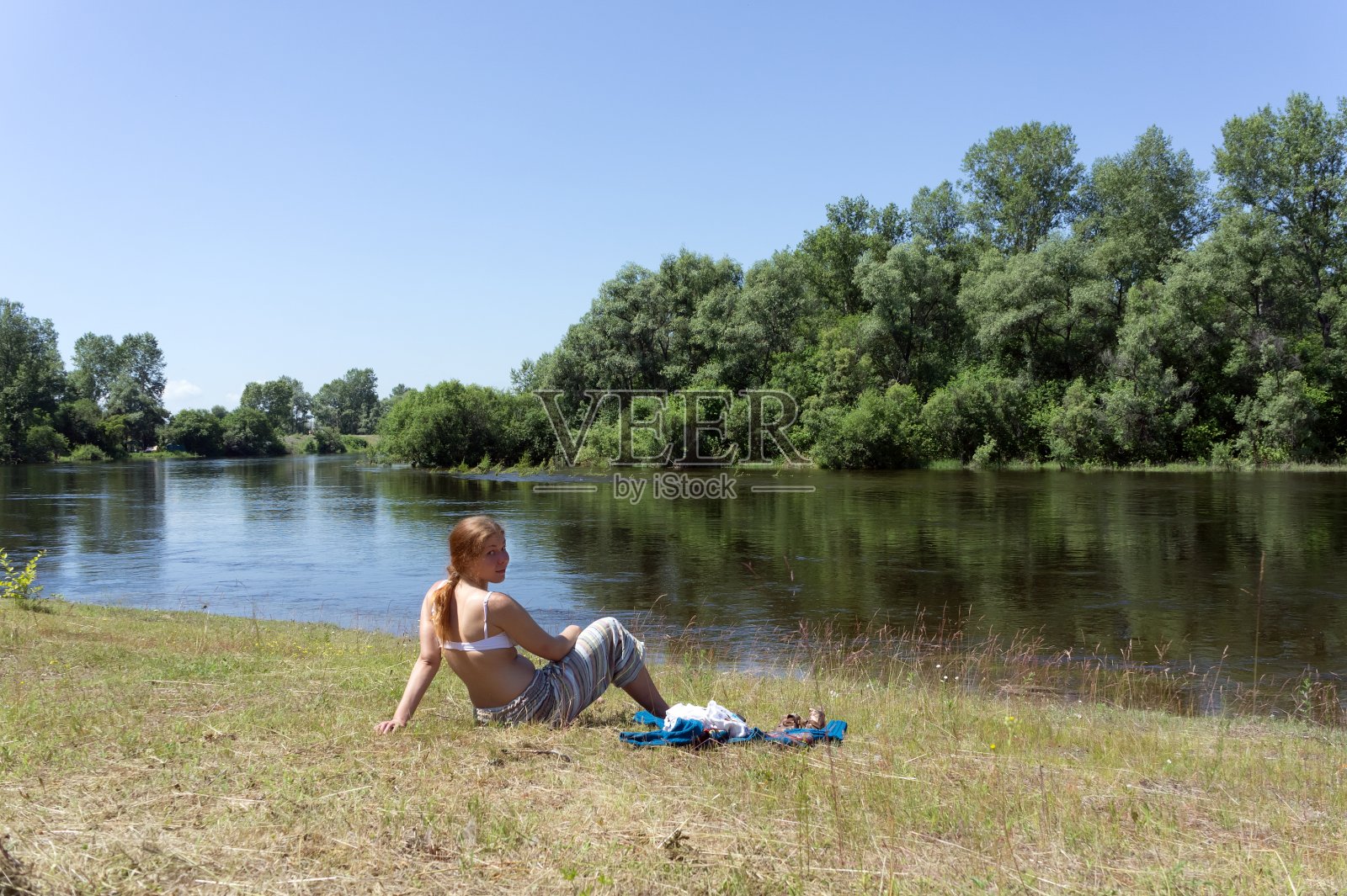 在一个阳光明媚的夏日，一个红头发的女孩坐在河边，背靠树林。照片摄影图片
