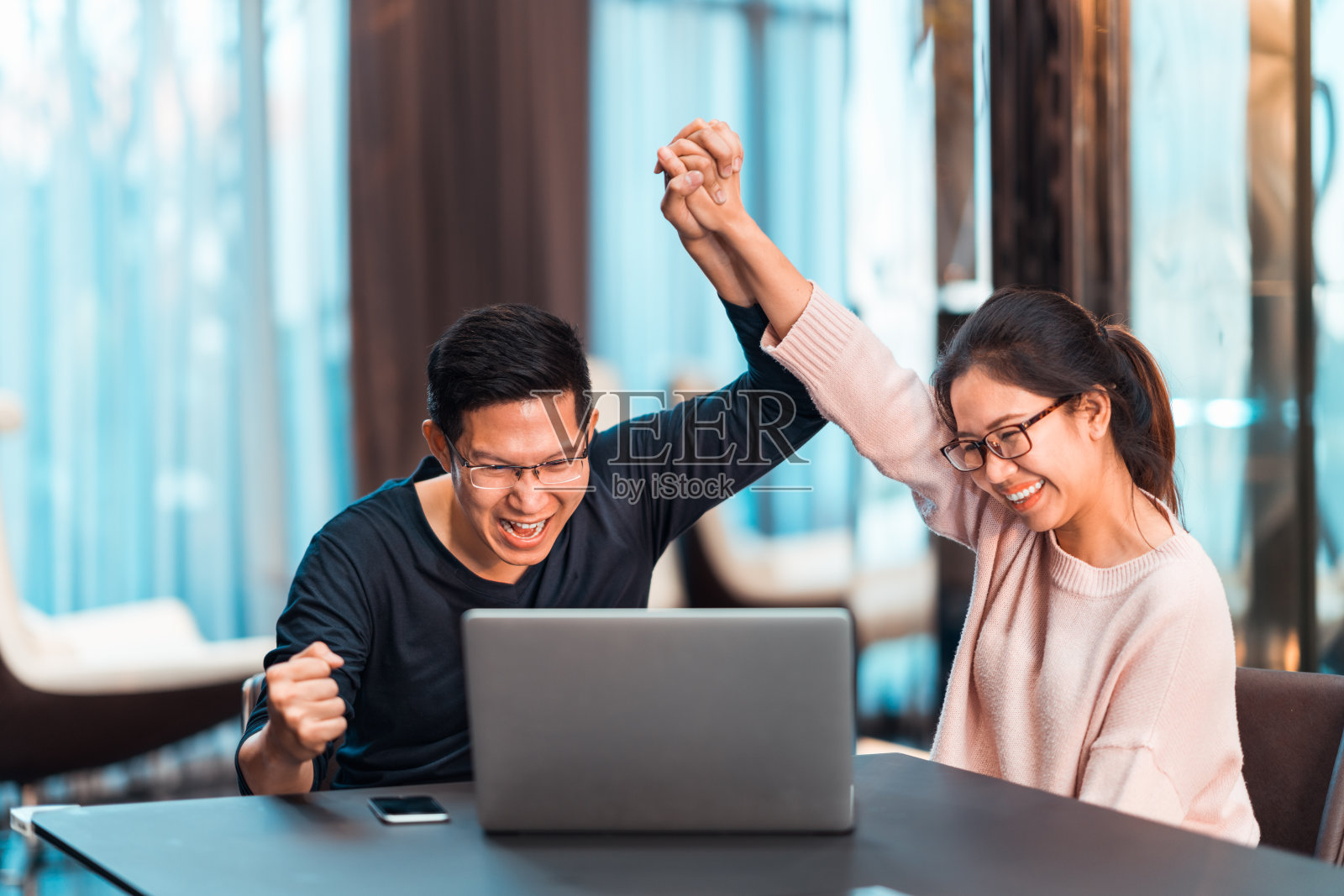 年轻的亚洲夫妇牵手一起庆祝，在现代家庭办公室看笔记本电脑。成功的工作，爱情关系，足球运动欢呼，或小型创业的概念。照片摄影图片