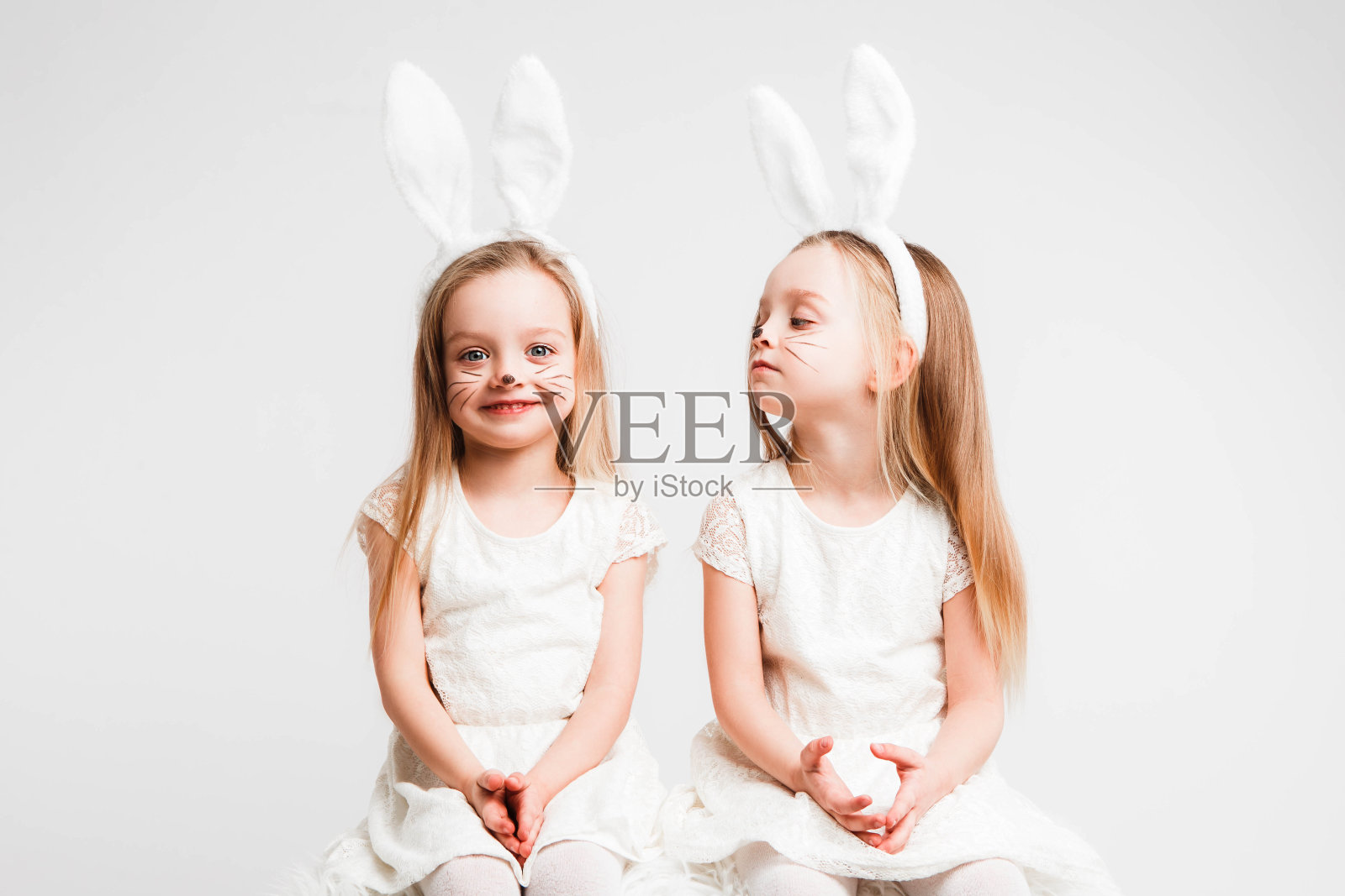 一对金发的双胞胎，穿着白裙子，长着兔耳朵。灰色背景上的工作室照片。孩子们庆祝复活节。照片摄影图片
