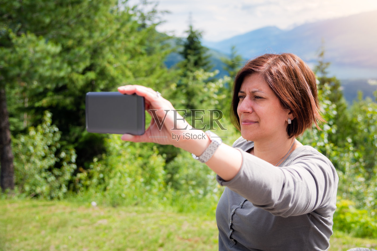 阳光明媚的一天，一名女游客在山顶用智能手机自拍照片摄影图片