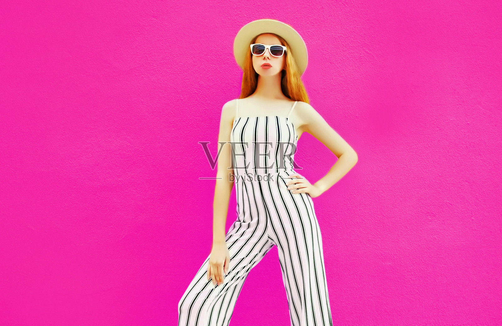 时尚的女人在夏天圆草帽，白色条纹连衣裤摆出彩色的粉红色墙壁背景照片摄影图片