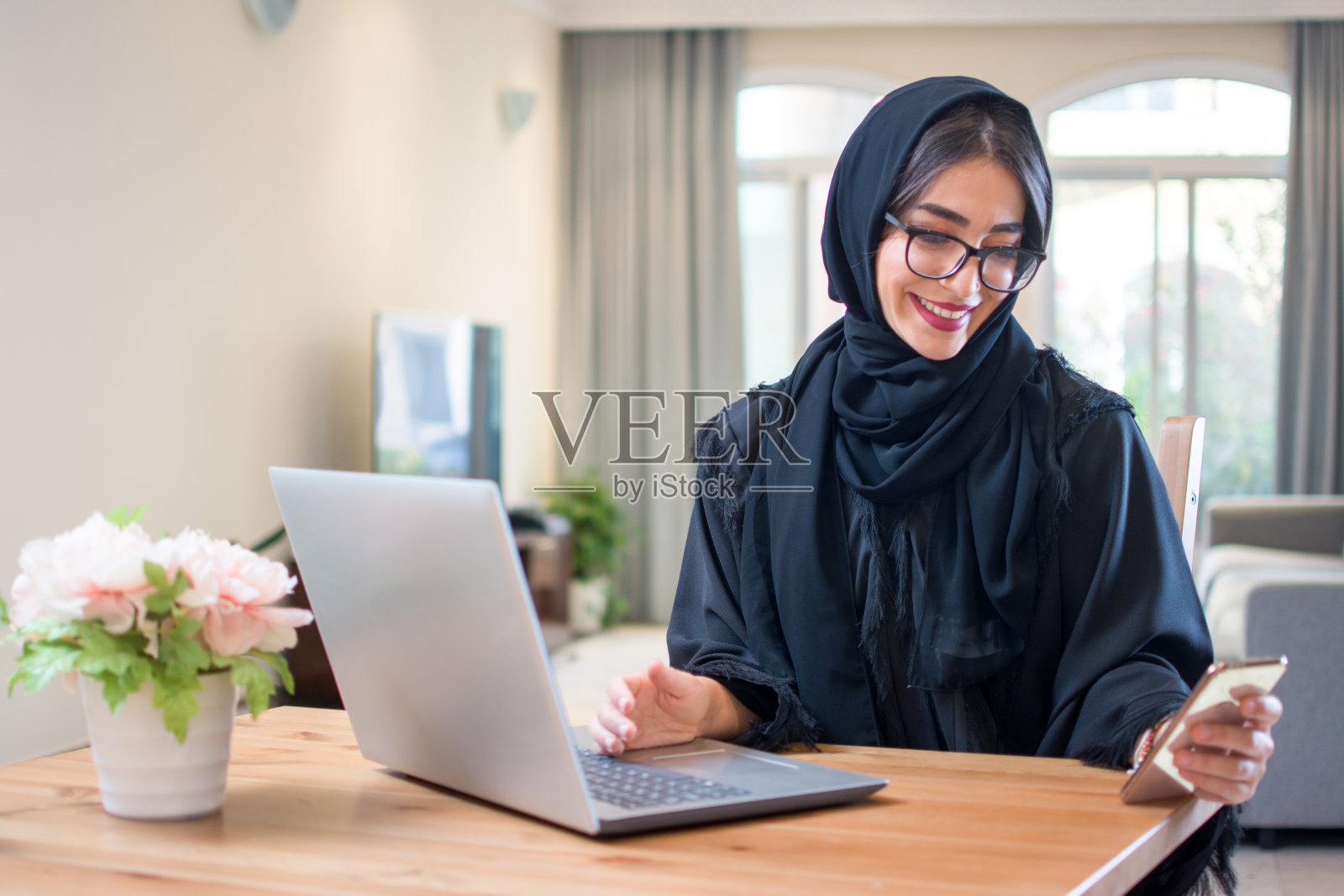 美丽快乐的穆斯林妇女在她的豪华家中使用智能手机和笔记本电脑照片摄影图片