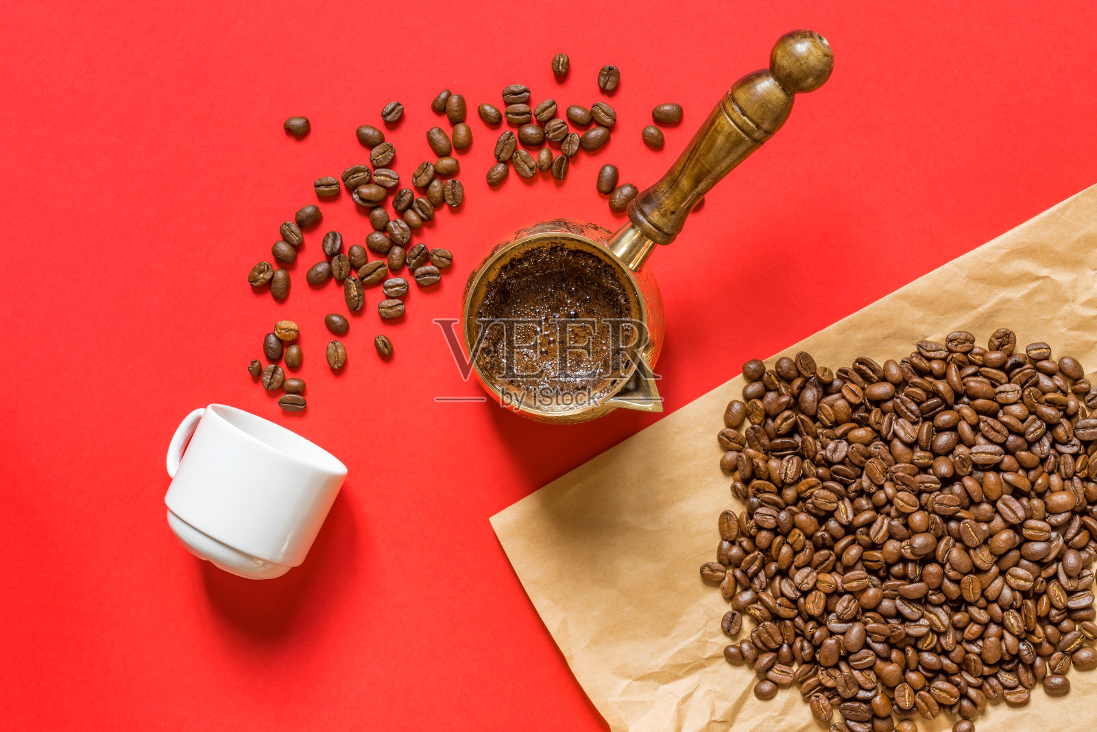 俯视图新鲜的咖啡在cezve(传统的土耳其咖啡壶)，白色的杯子和咖啡豆在牛皮纸在红色的背景。照片摄影图片