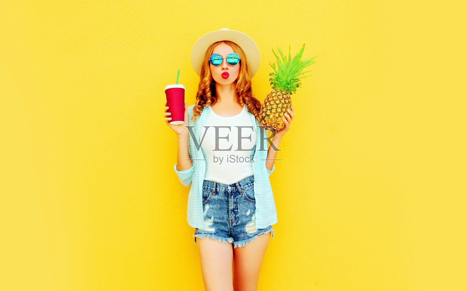 肖像时尚的女人送甜蜜的飞吻菠萝，一杯果汁在夏季草帽，太阳镜，短裤在彩色的黄色背景照片摄影图片