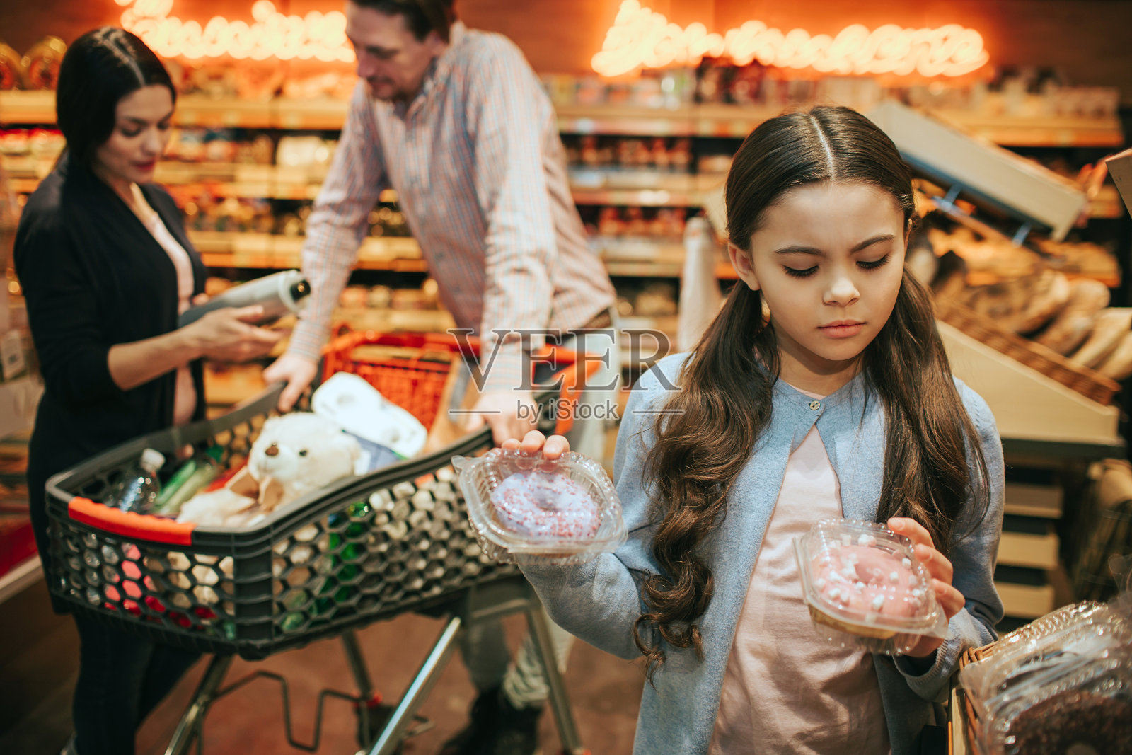 年轻的父母和女儿在杂货店里。女孩拿着两个装有甜甜圈的盒子，看着它们。父母站在购物车后面。照片摄影图片