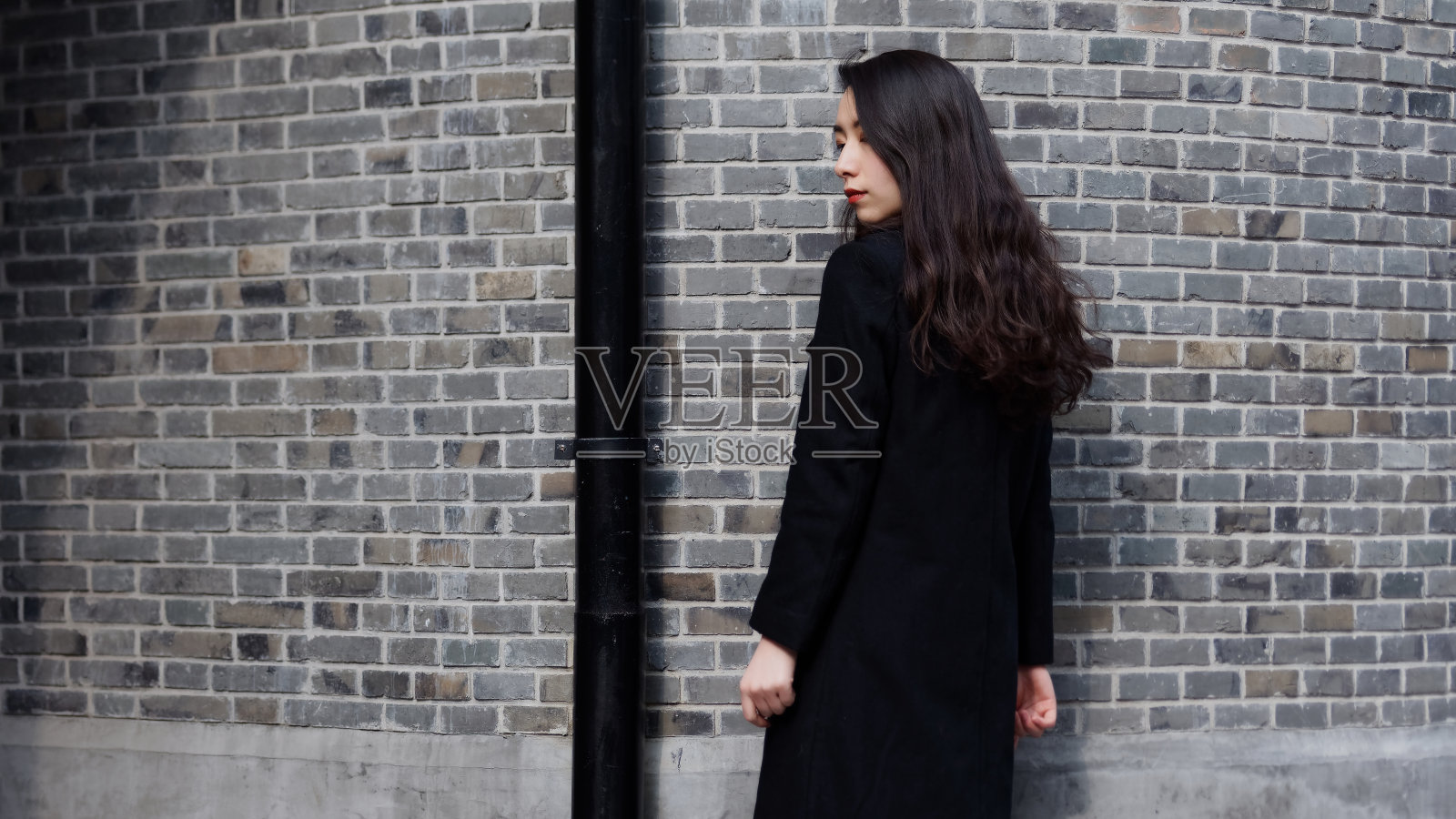 美丽的中国年轻女子穿着黑色外套在复古砖墙前摆姿势，侧视图，户外时尚写真魅力时尚女士。照片摄影图片