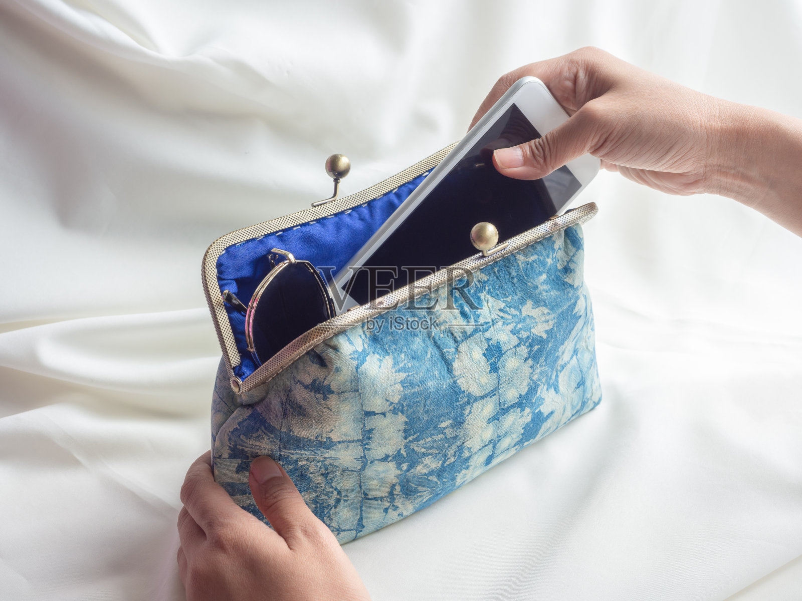 女人的手把白色的智能手机和太阳镜放在复古的靛蓝布钱包里。照片摄影图片