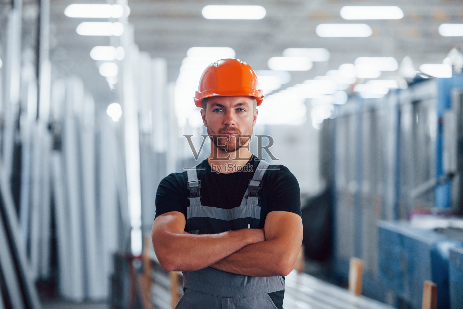 双臂交叉站着。男性工业工人在工厂室内的肖像。戴着橙色安全帽的年轻技术员照片摄影图片