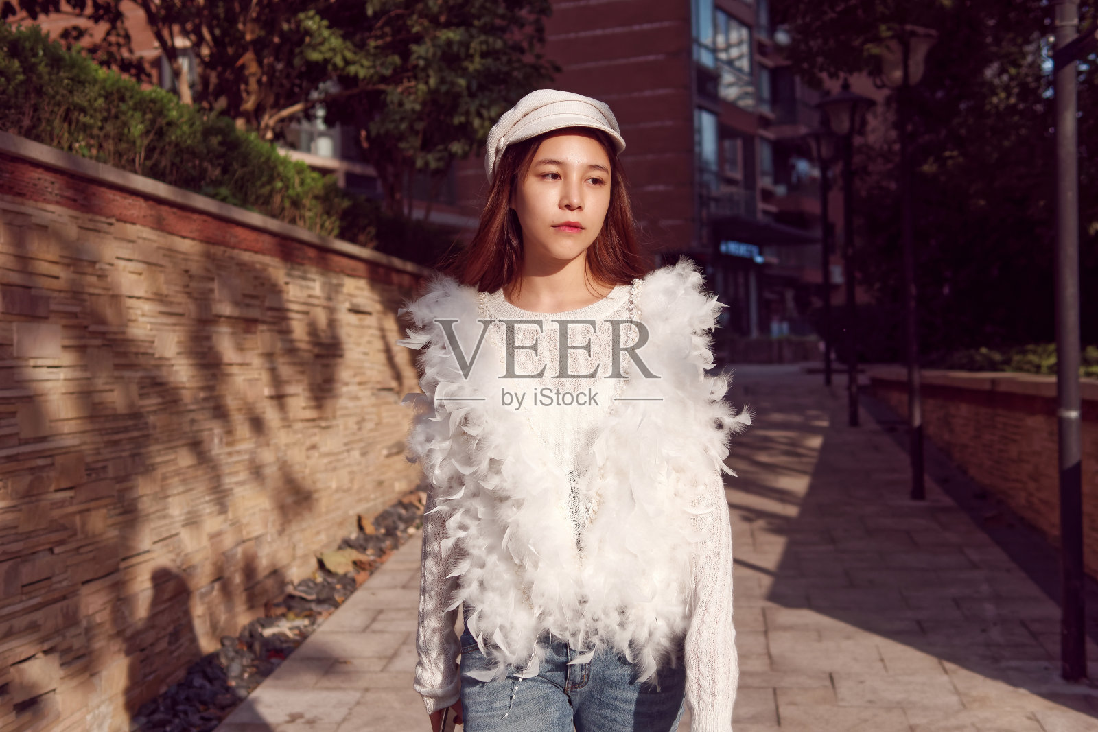 一个美丽的中国年轻女子的肖像，穿着白色羽毛衣服和牛仔裤走在阳光灿烂的街道上，漫无目的地看着别处。照片摄影图片
