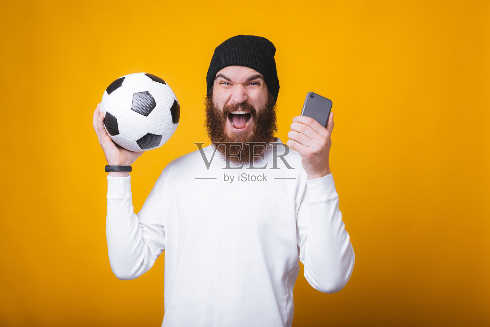 一个年轻人正尖叫着，手里拿着一个电话和一个足球靠近黄色的墙。照片摄影图片