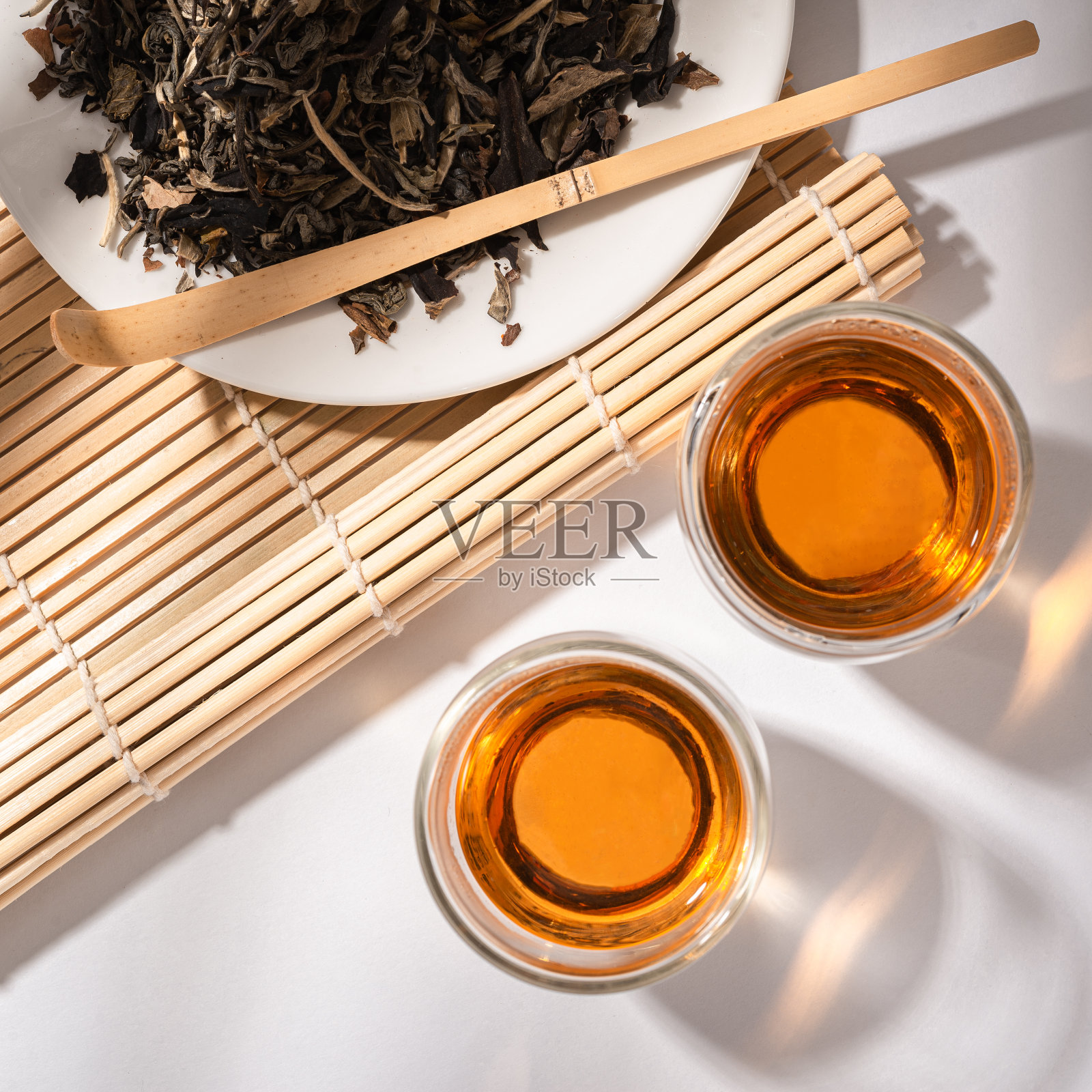 新鲜冲泡的茶在玻璃杯中，干绿茶叶子在一个白色的盘子，竹配件冲泡仪式，树荫的背景照片摄影图片