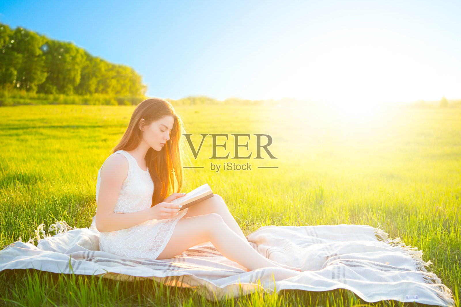 在一个阳光明媚的日子里，一个女孩坐在格子裙上，在绿草如茵的田野里看书照片摄影图片