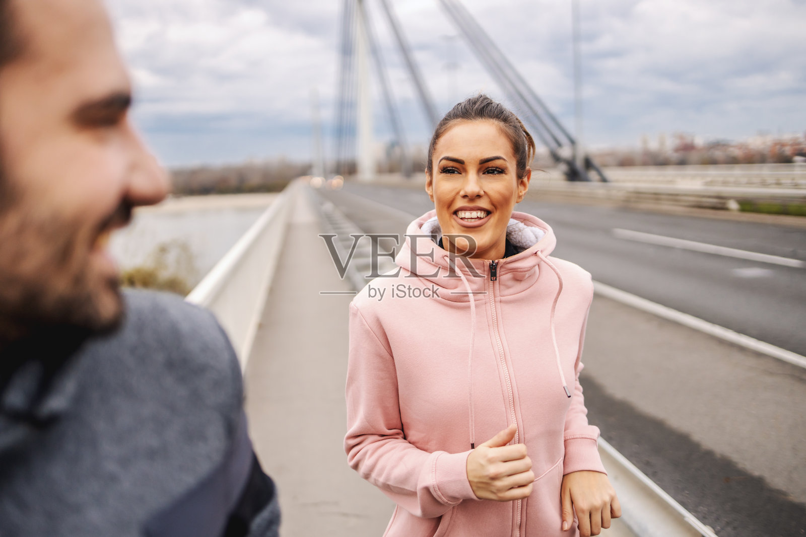 近距离的健身女运动员穿着运动服在桥上跑步和她的男性朋友看他。健身户外多云天气的概念。照片摄影图片