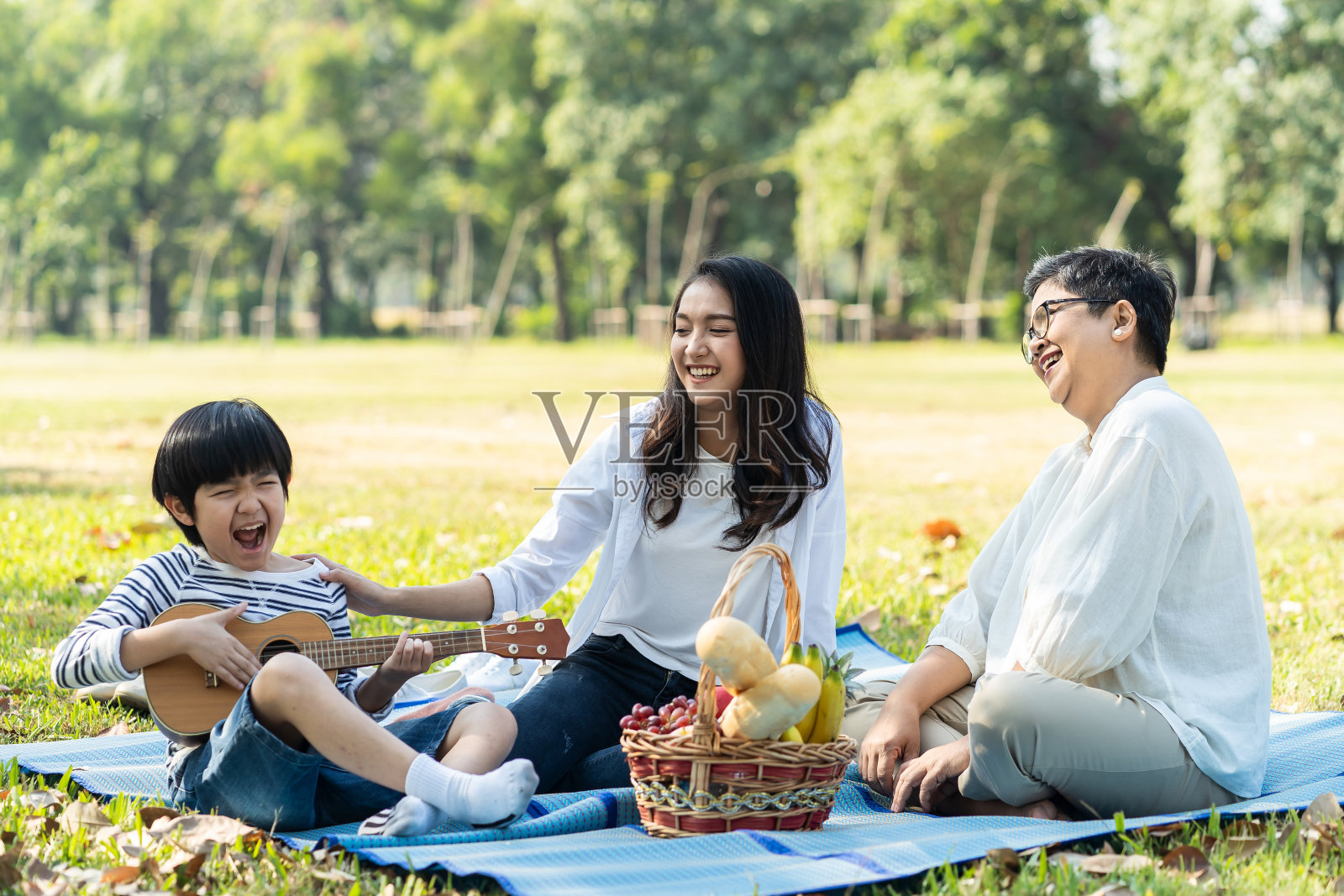 亚洲家庭在新鲜的绿色公园一起野餐。妈妈微笑着抚摸孩子，奶奶开心地看着孩子弹尤克里里或迷你吉他。放松的家庭活动。照片摄影图片