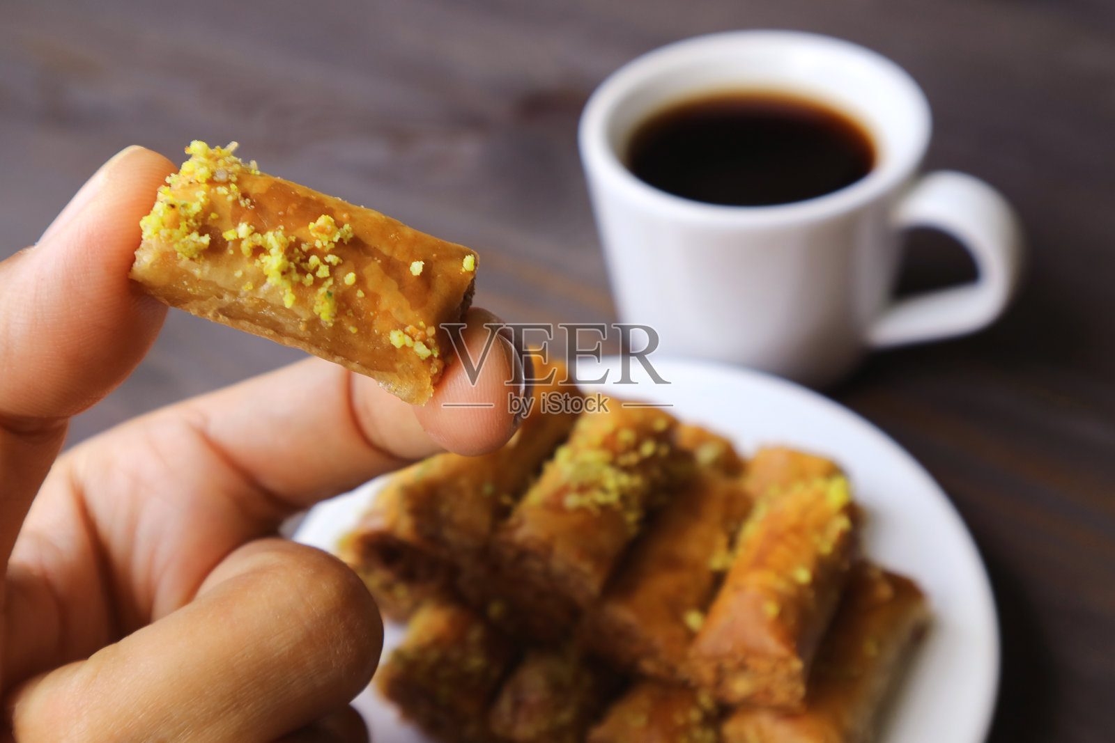 手指拿着开心果果仁酥饼和模糊的土耳其咖啡和一盘糖果在背景照片摄影图片