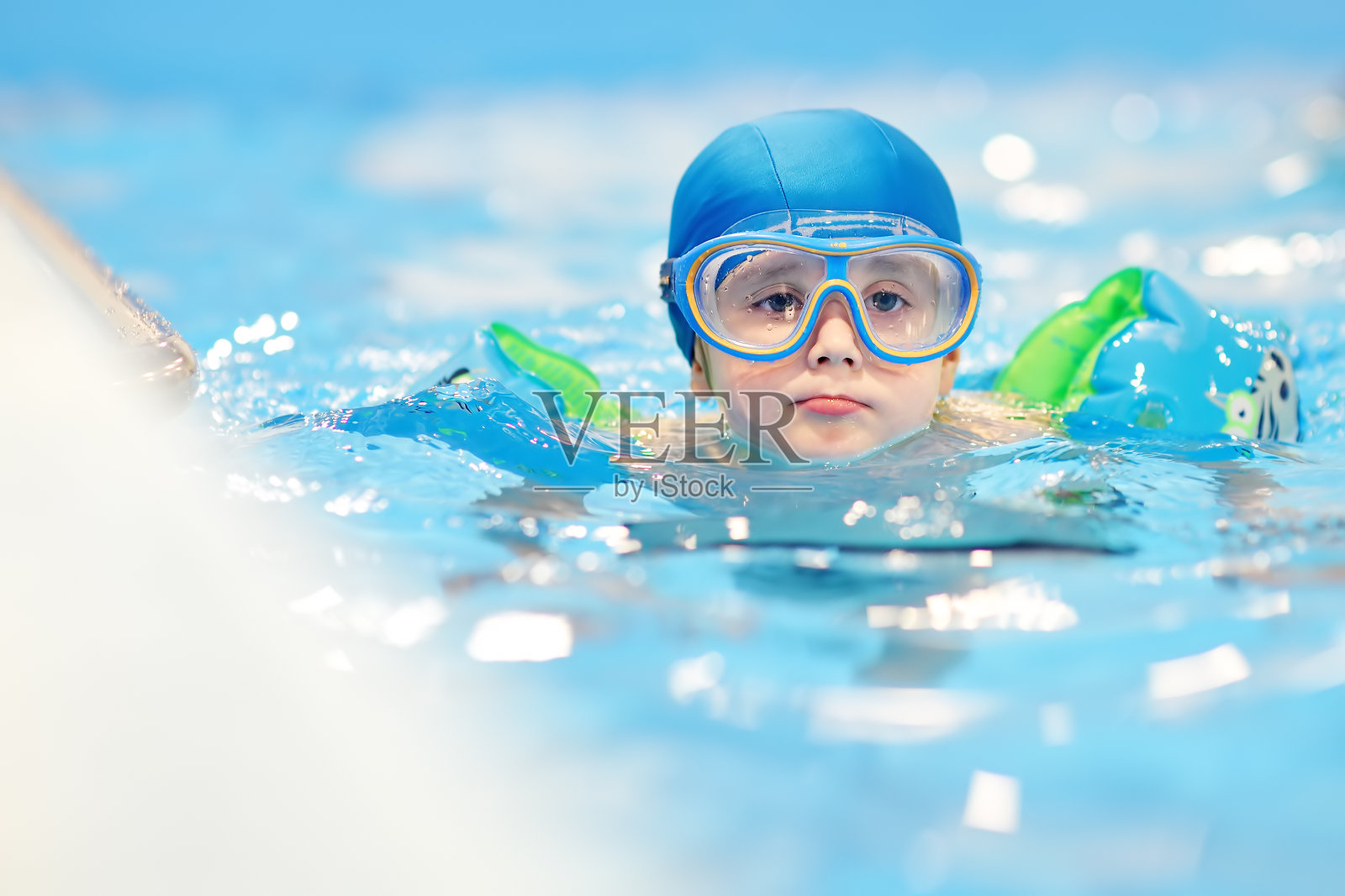 戴着眼镜戴着充气臂圈的小男孩在游泳池里。照片摄影图片
