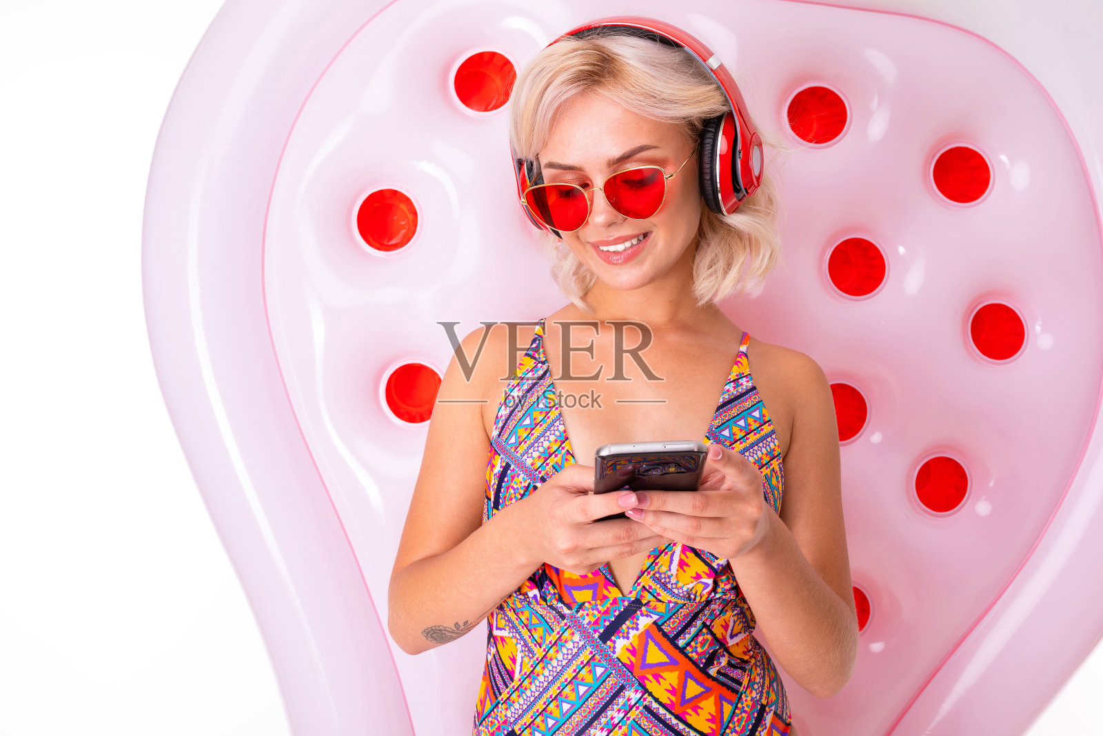 性感的金发女孩穿着泳衣，戴着太阳镜，手里拿着手机，背景是一个游泳床垫，她用耳机听音乐照片摄影图片
